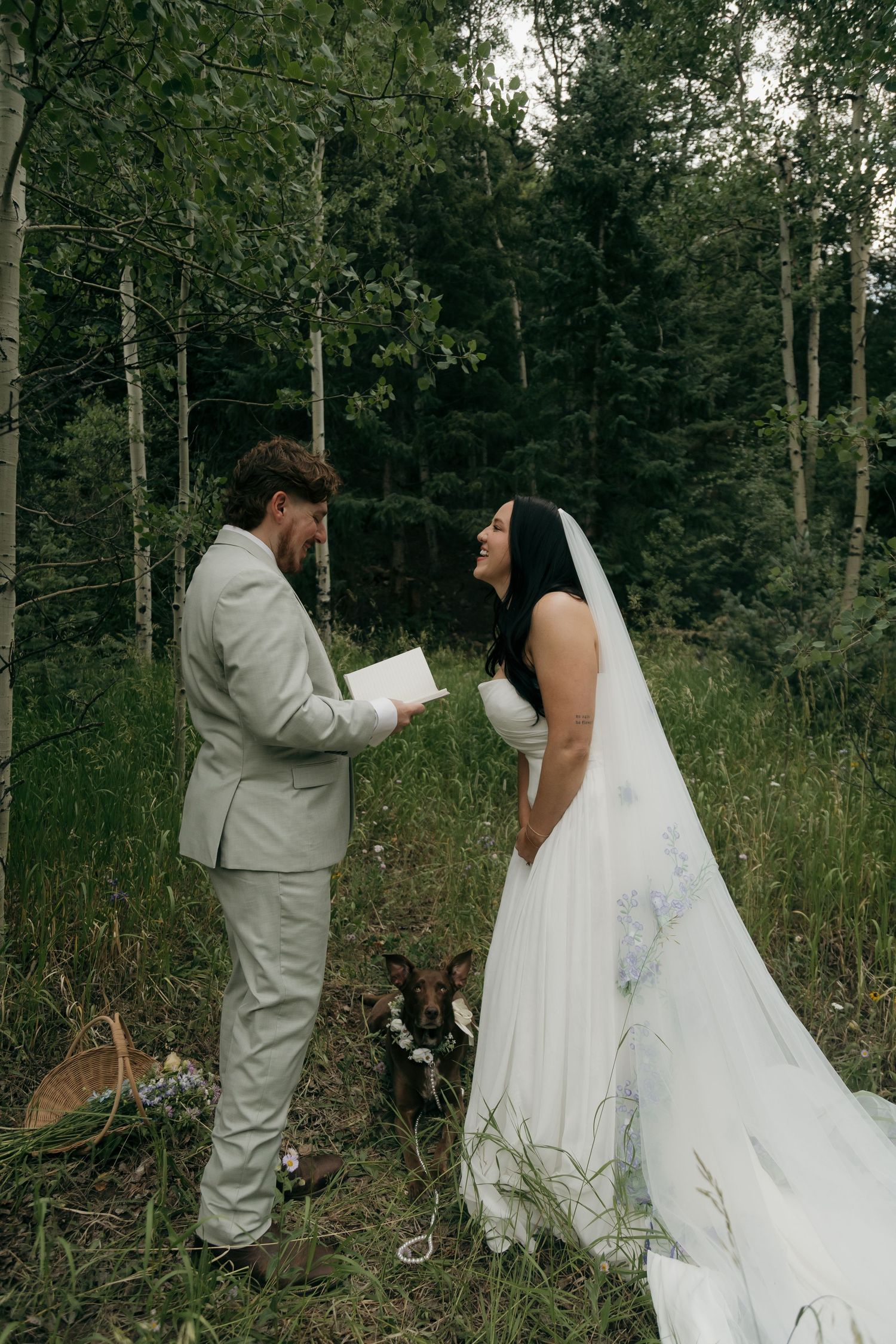 Bride and groom exchange vows in a lush forest setting, with a dog sitting between them during their outdoor wedding ceremony.