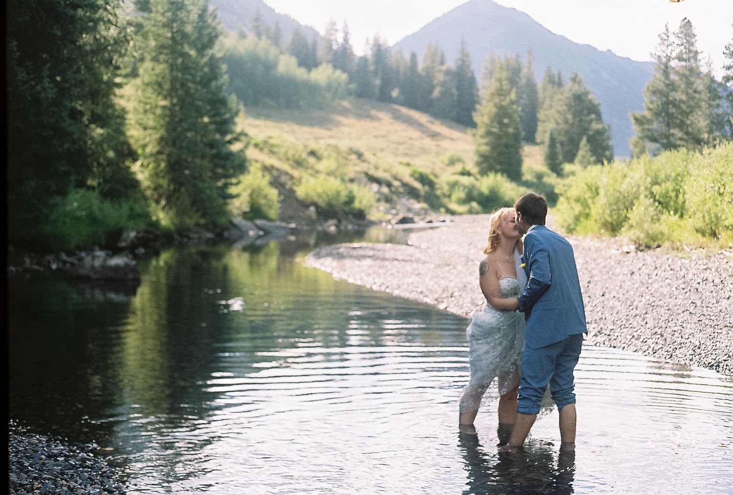 Bride and groom stand kissing in a mountain stream surrounded by lush green trees and hills.