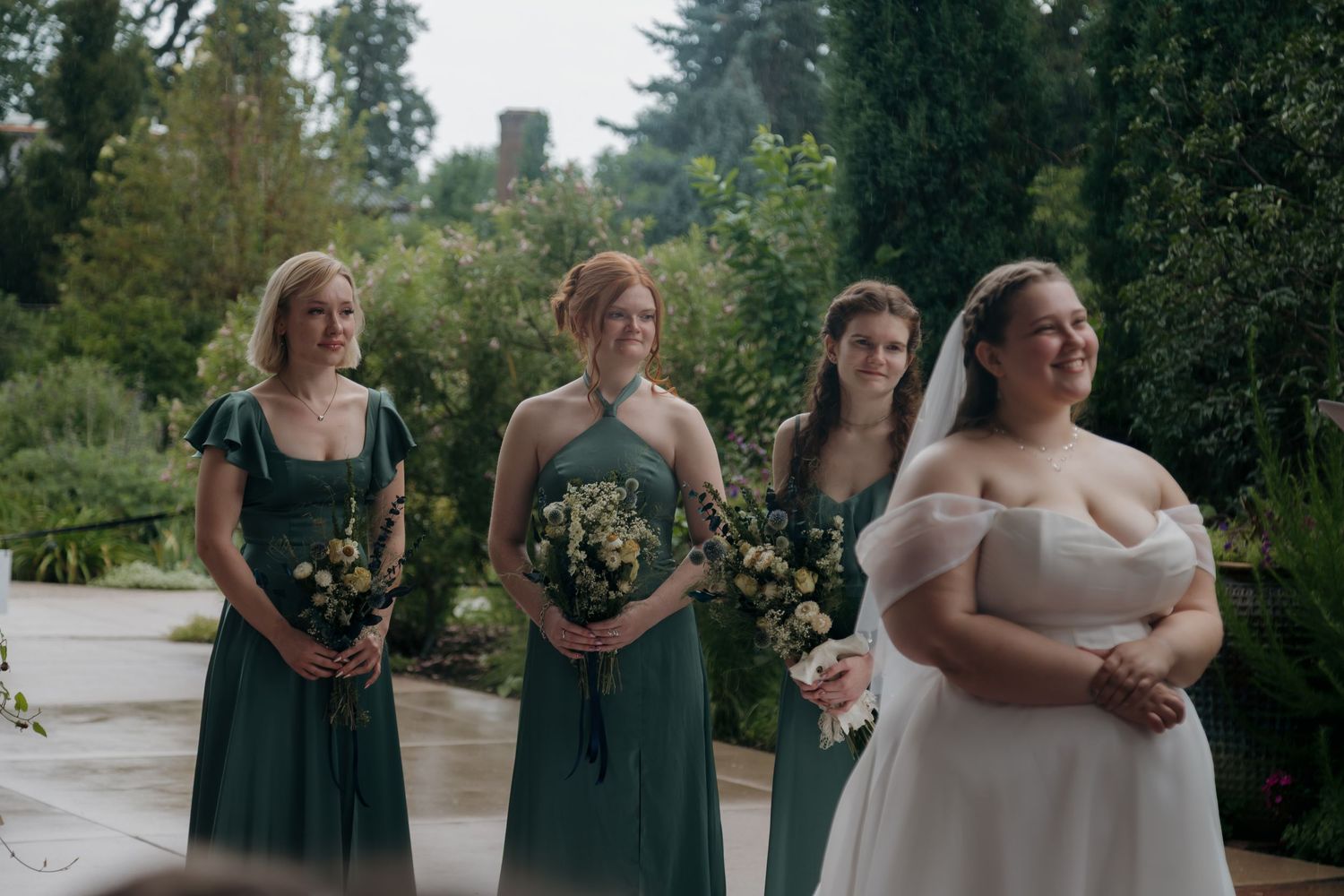Bride in white off-shoulder gown stands with three bridesmaids in green dresses holding bouquets.