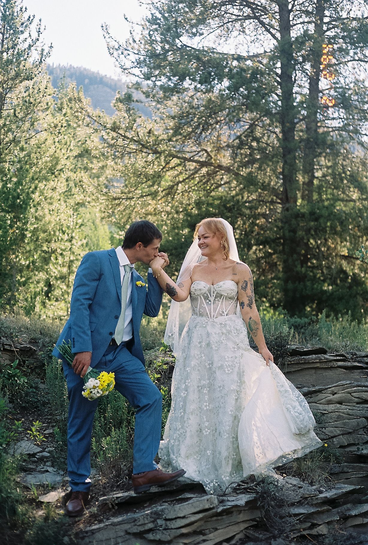 Groom in blue suit kisses bride's hand outdoors, bride wearing lace wedding dress with veil, forest backdrop.