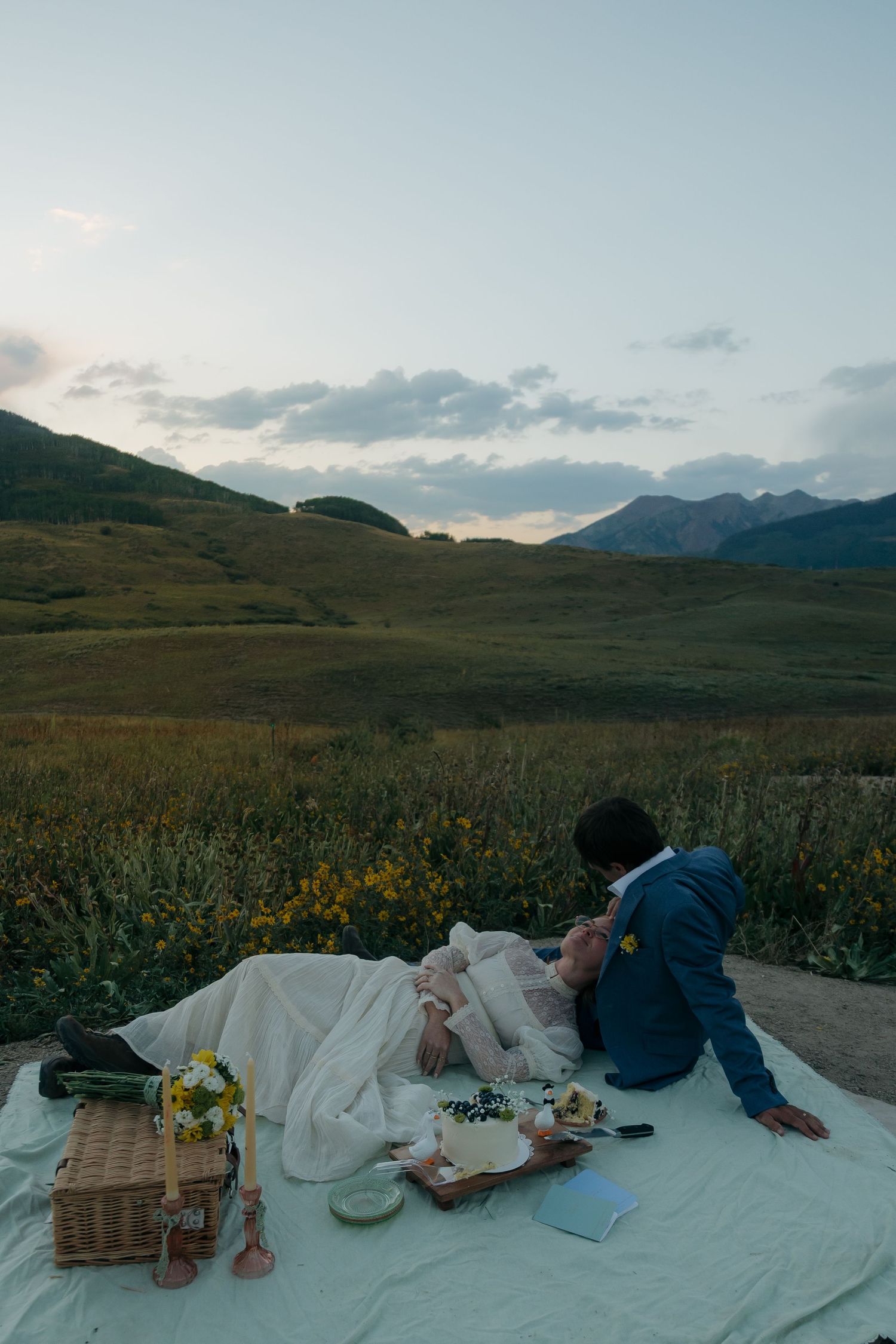 Bride and groom enjoying romantic picnic on white blanket in golden mountain meadow at dusk.