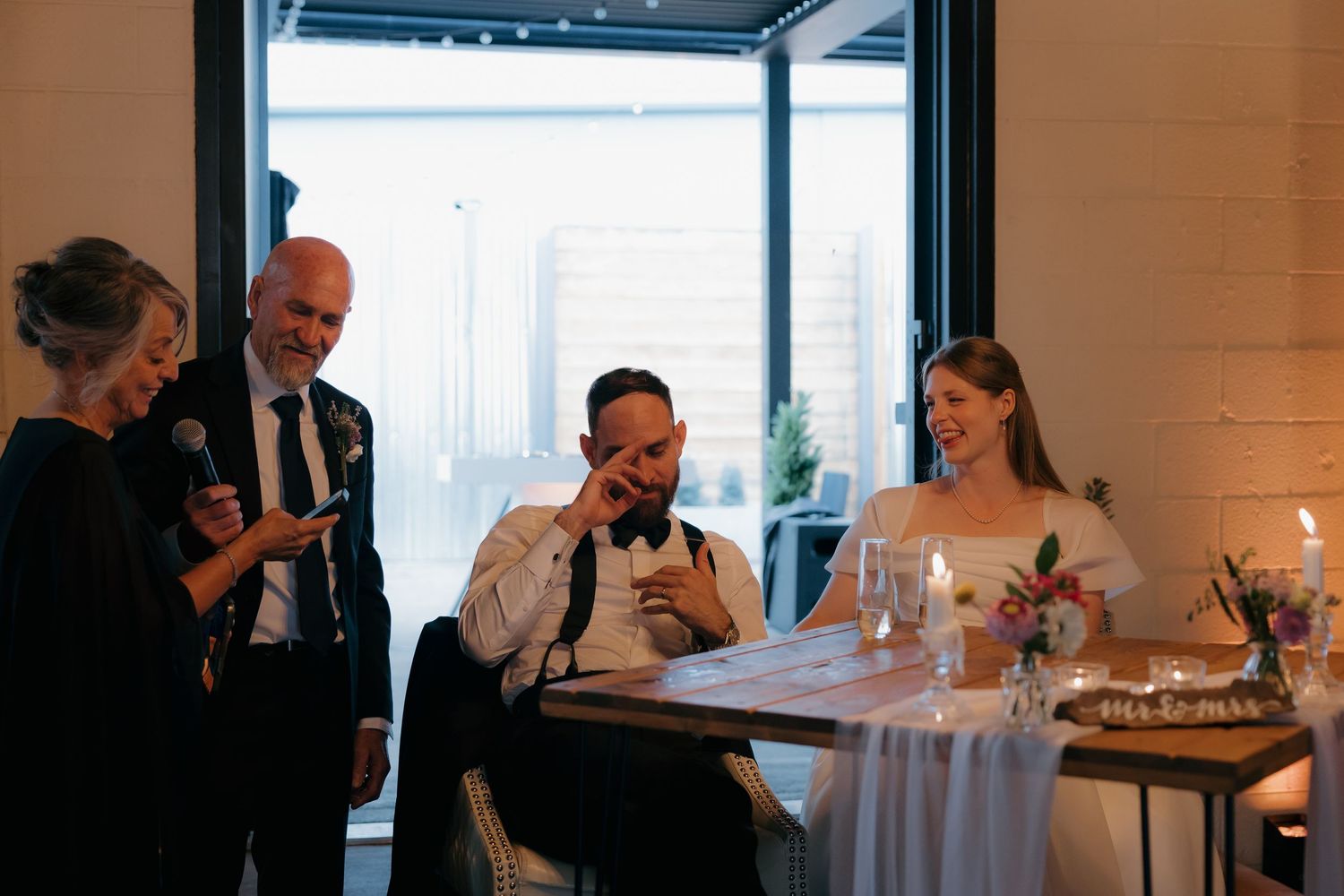 Parents delivering heartfelt wedding speech to laughing bride and groom at reception table.