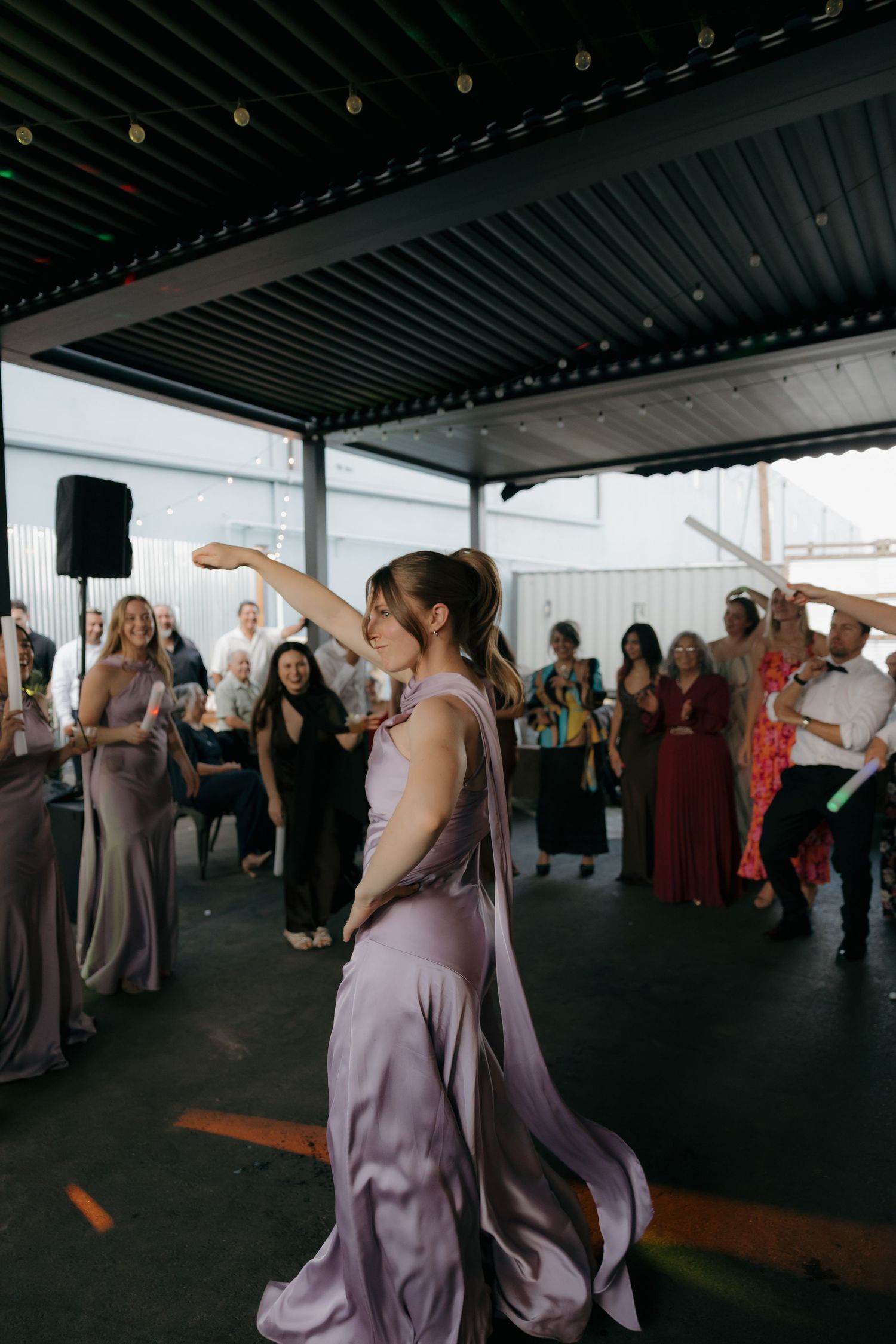 Bridesmaid in lavender satin gown twirls gracefully on dance floor while wedding guests cheer at outdoor reception.