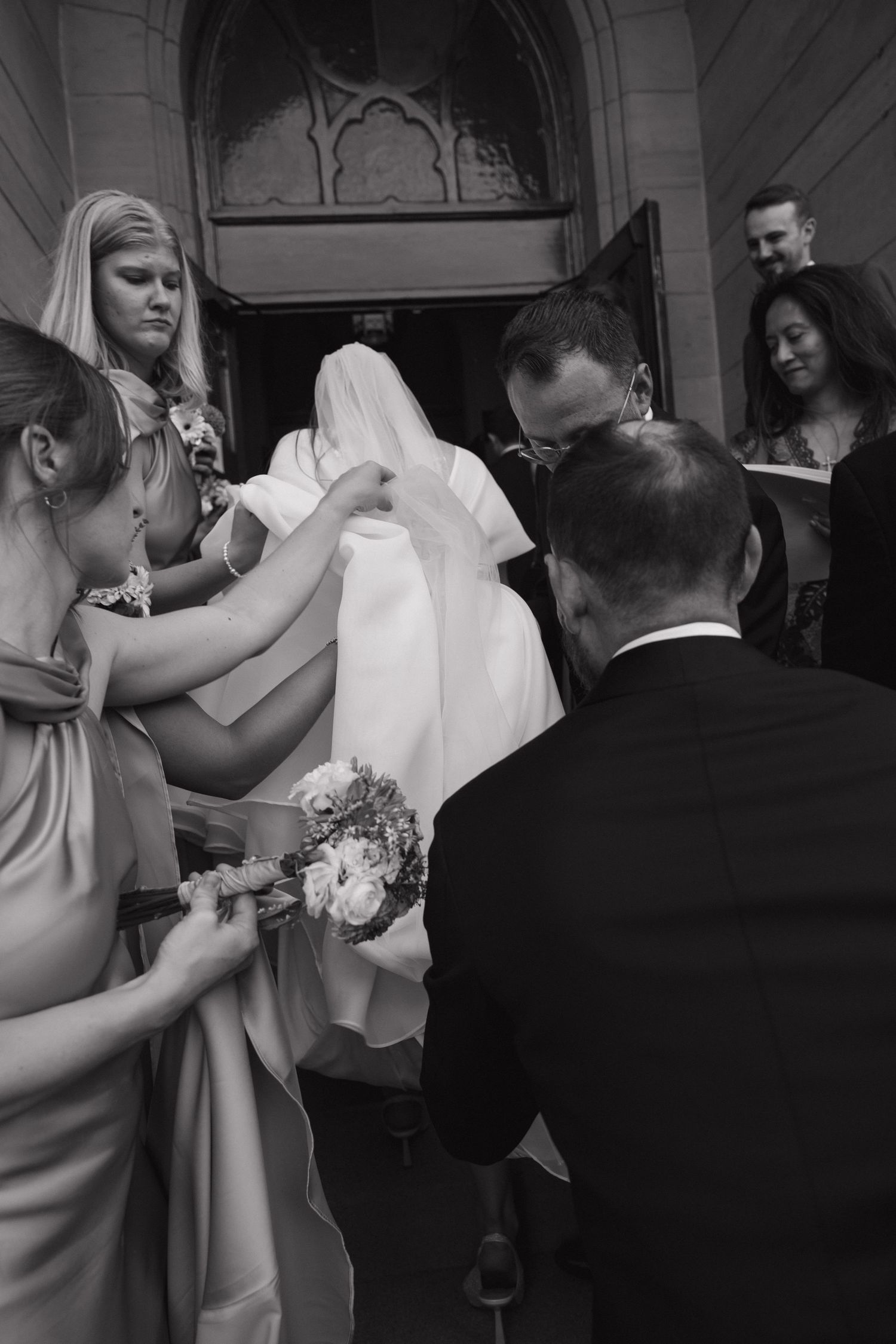 Black and white photo of bridesmaids adjusting bride's veil and gown train at church entrance before ceremony.