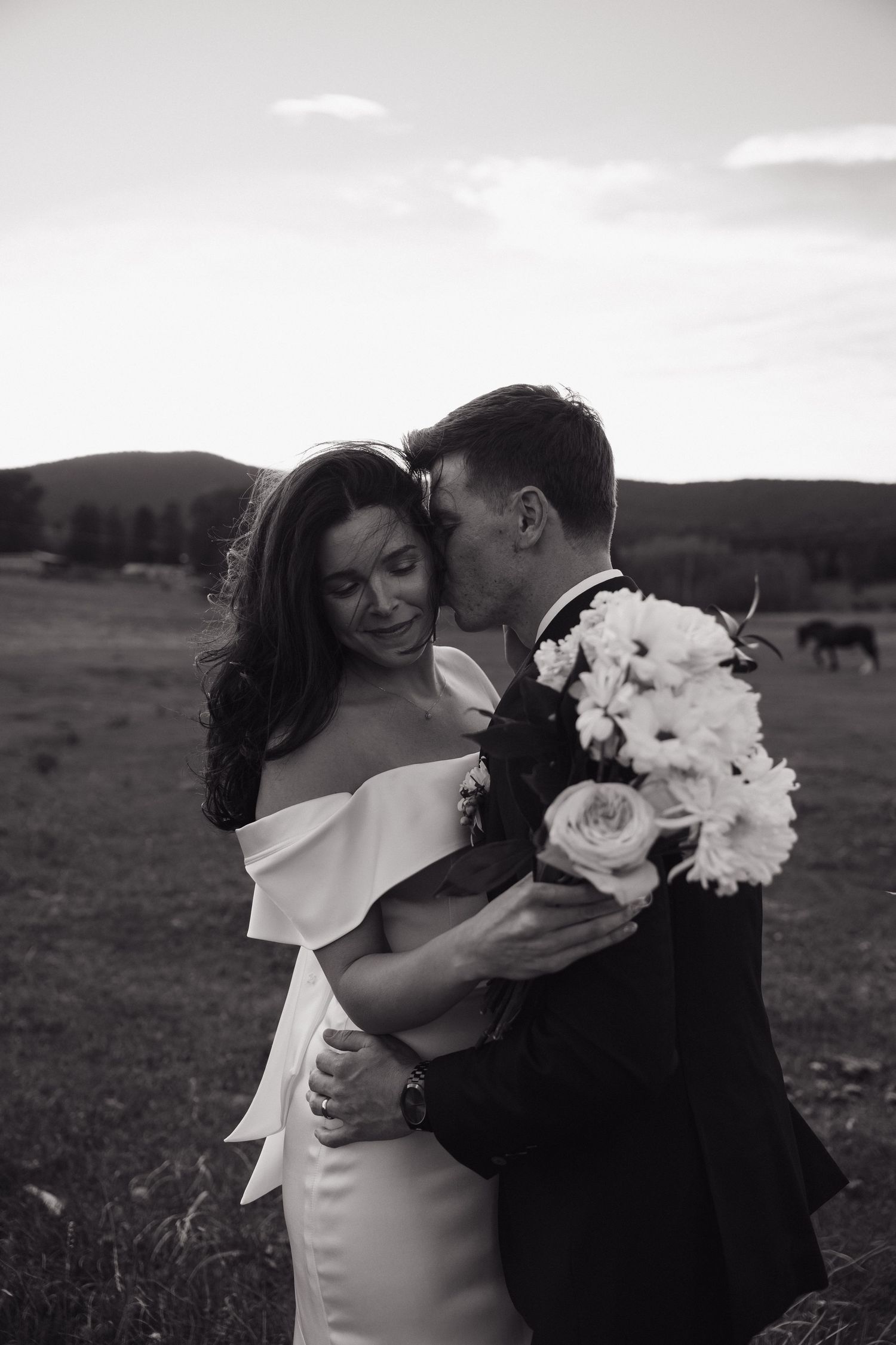 Black and white photo of a groom kissing bride's cheek while holding a bouquet in a scenic field.