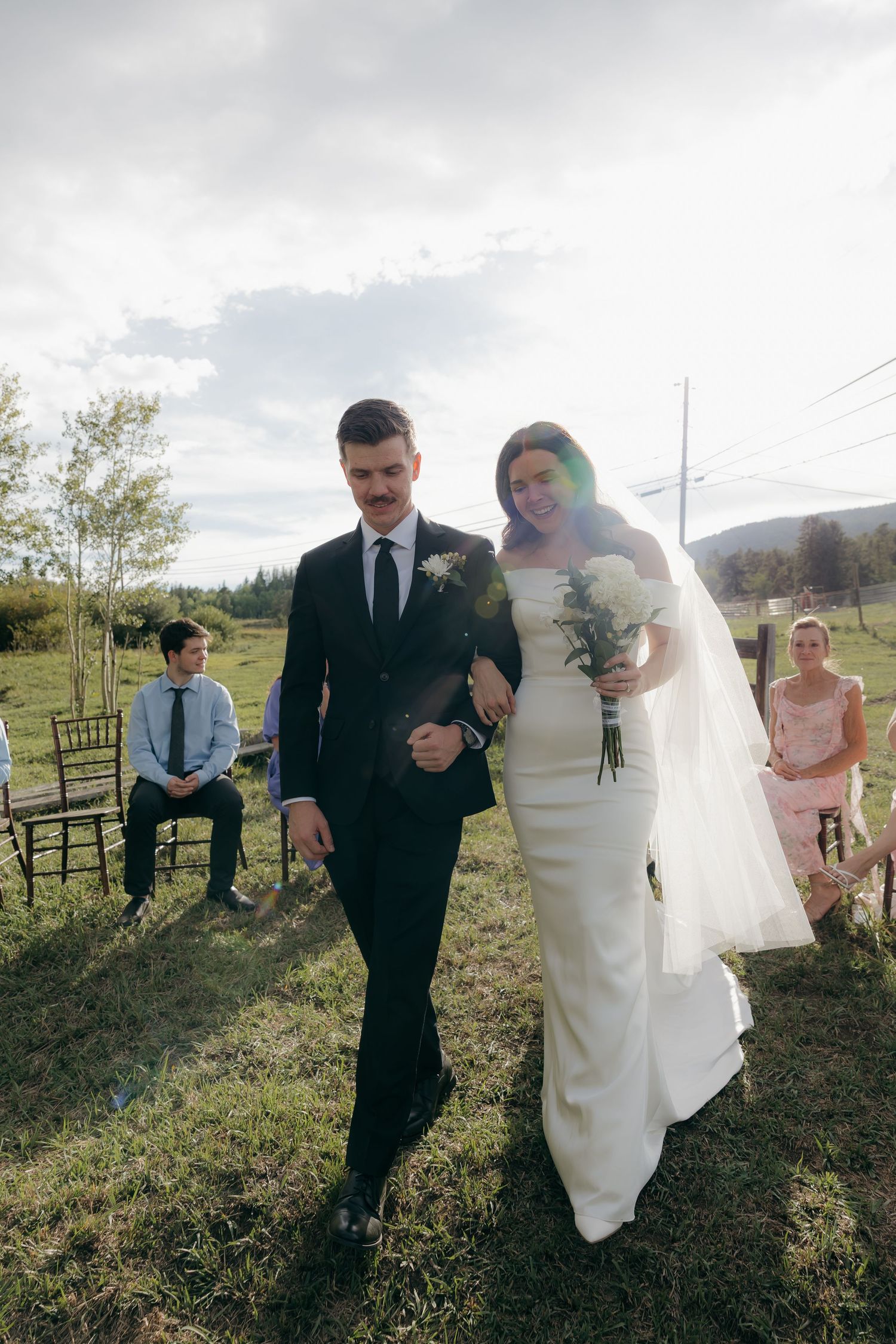 Bride in white gown and groom in black suit walk arm-in-arm down outdoor aisle at rustic countryside wedding ceremony.