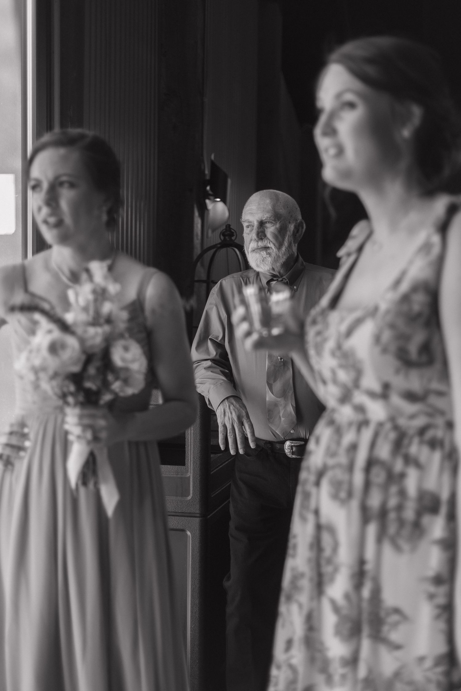 Black and white wedding photo of bridesmaids holding bouquet and drink with elderly man standing in background.