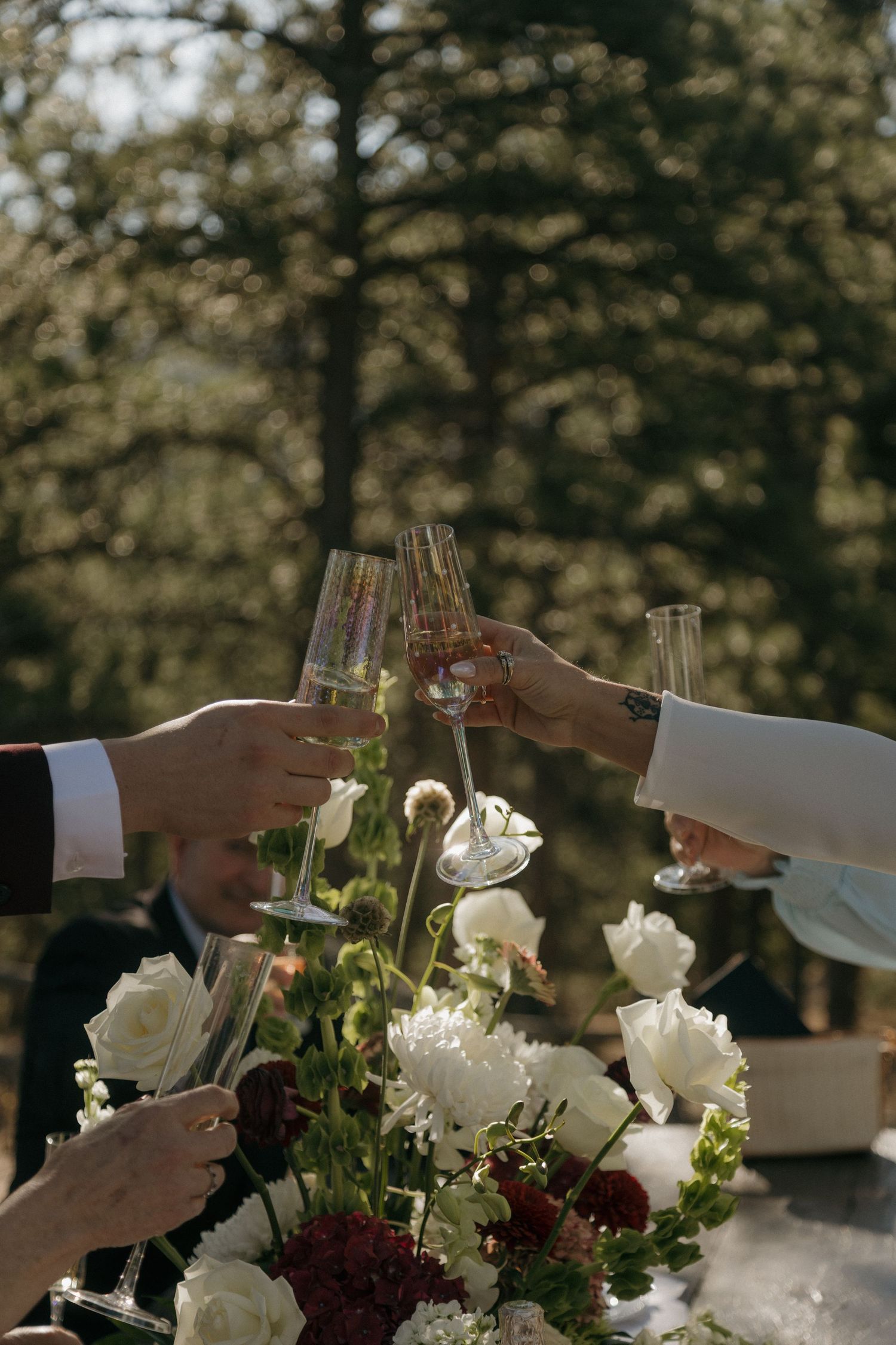 Three hands raise wine glasses in a toast over an elegant table setting with white roses and burgundy flowers.