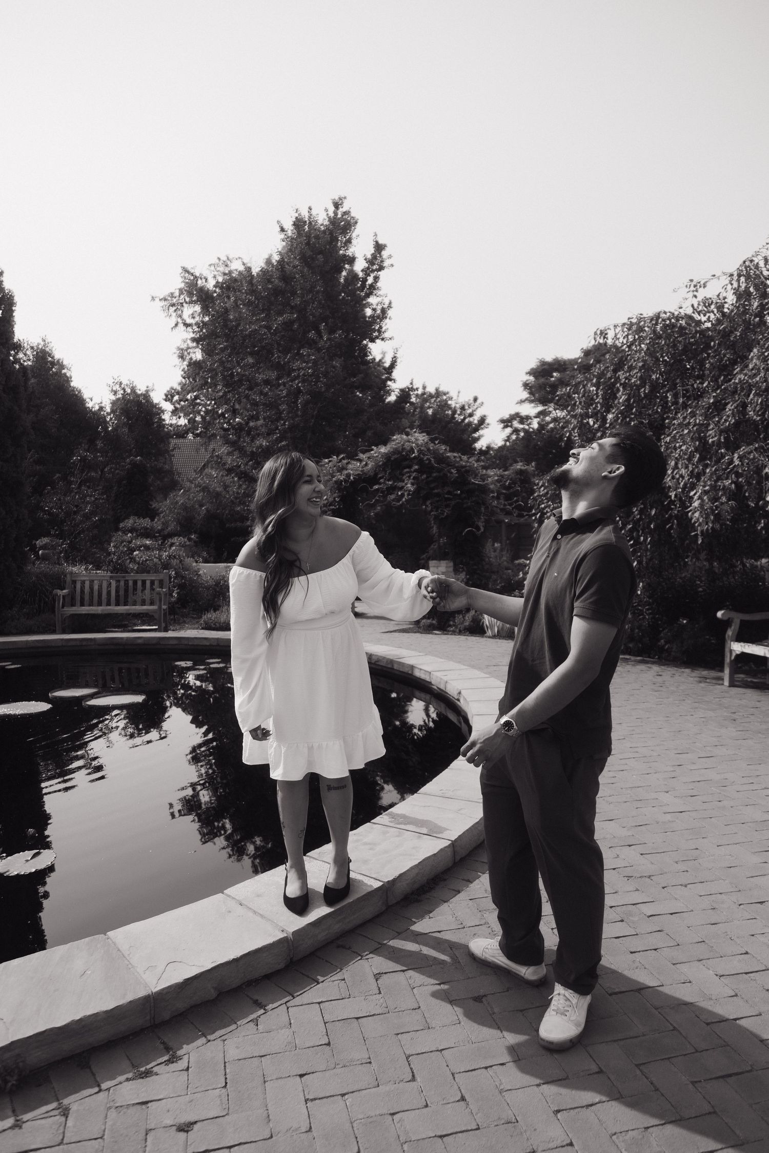 Black and white photo of a couple holding hands by a garden pond, woman in white off-shoulder dress smiling at man.