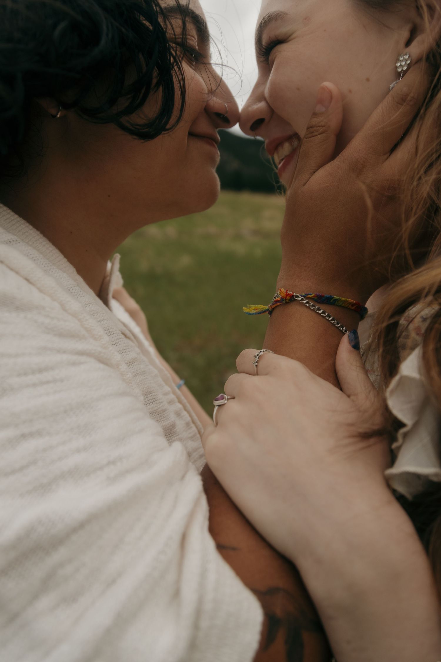 Two women share an intimate nose-to-nose moment, smiling joyfully, wearing colorful bracelets outdoors.