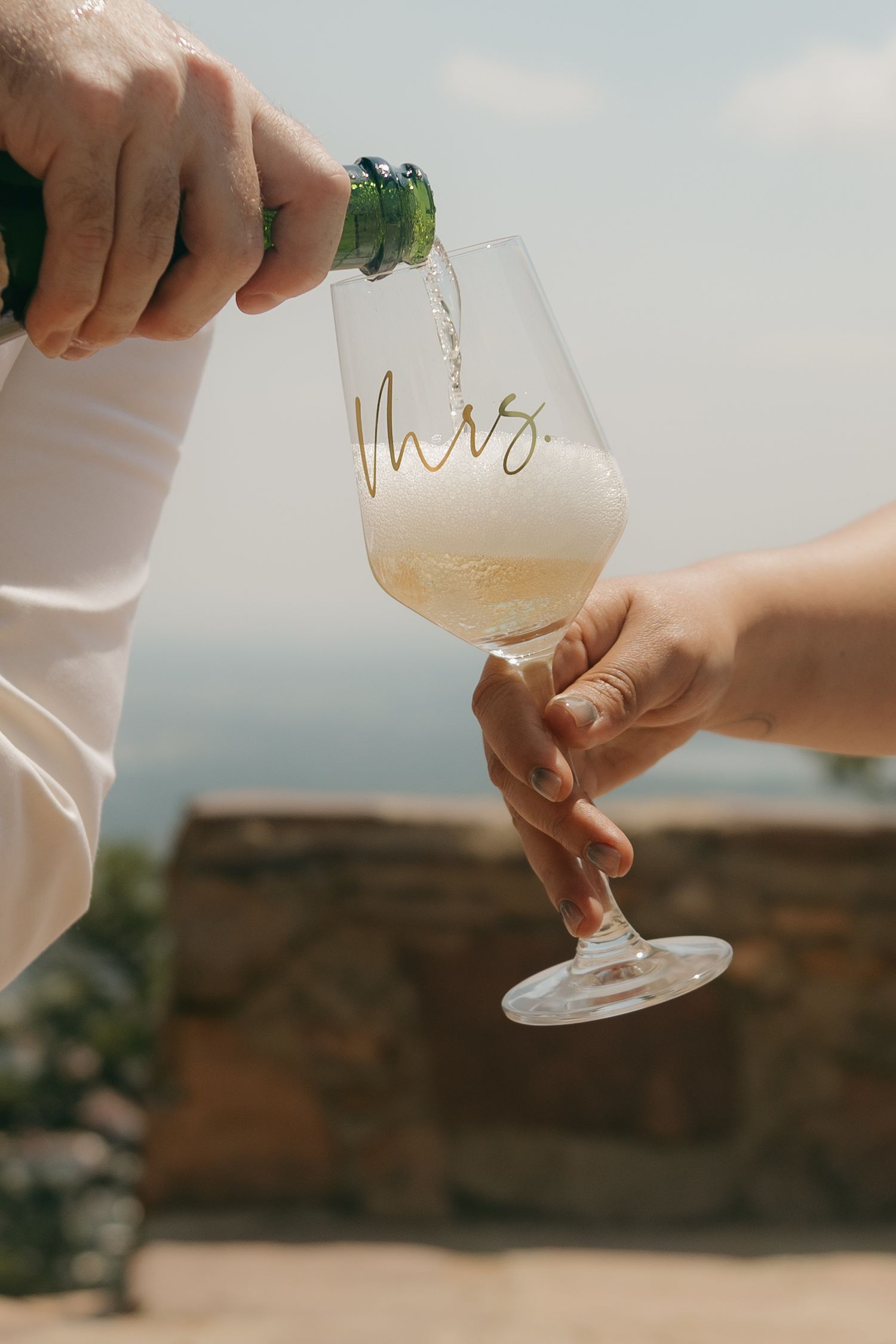 Champagne being poured into a crystal glass etched with Mrs. in gold script at an outdoor wedding celebration.