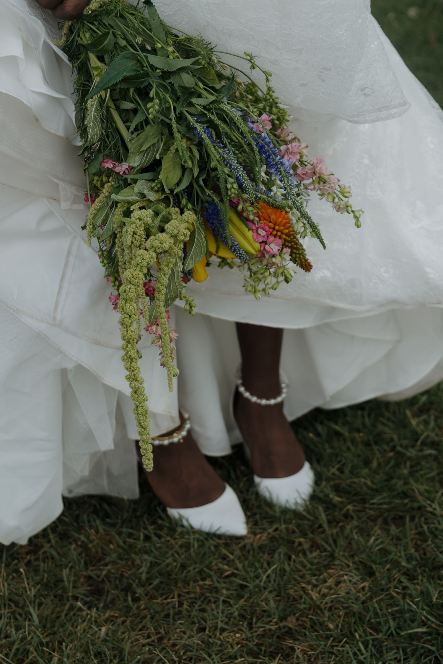 Bride in white gown holding lush wildflower bouquet wearing white heels with pearl anklets on grass.