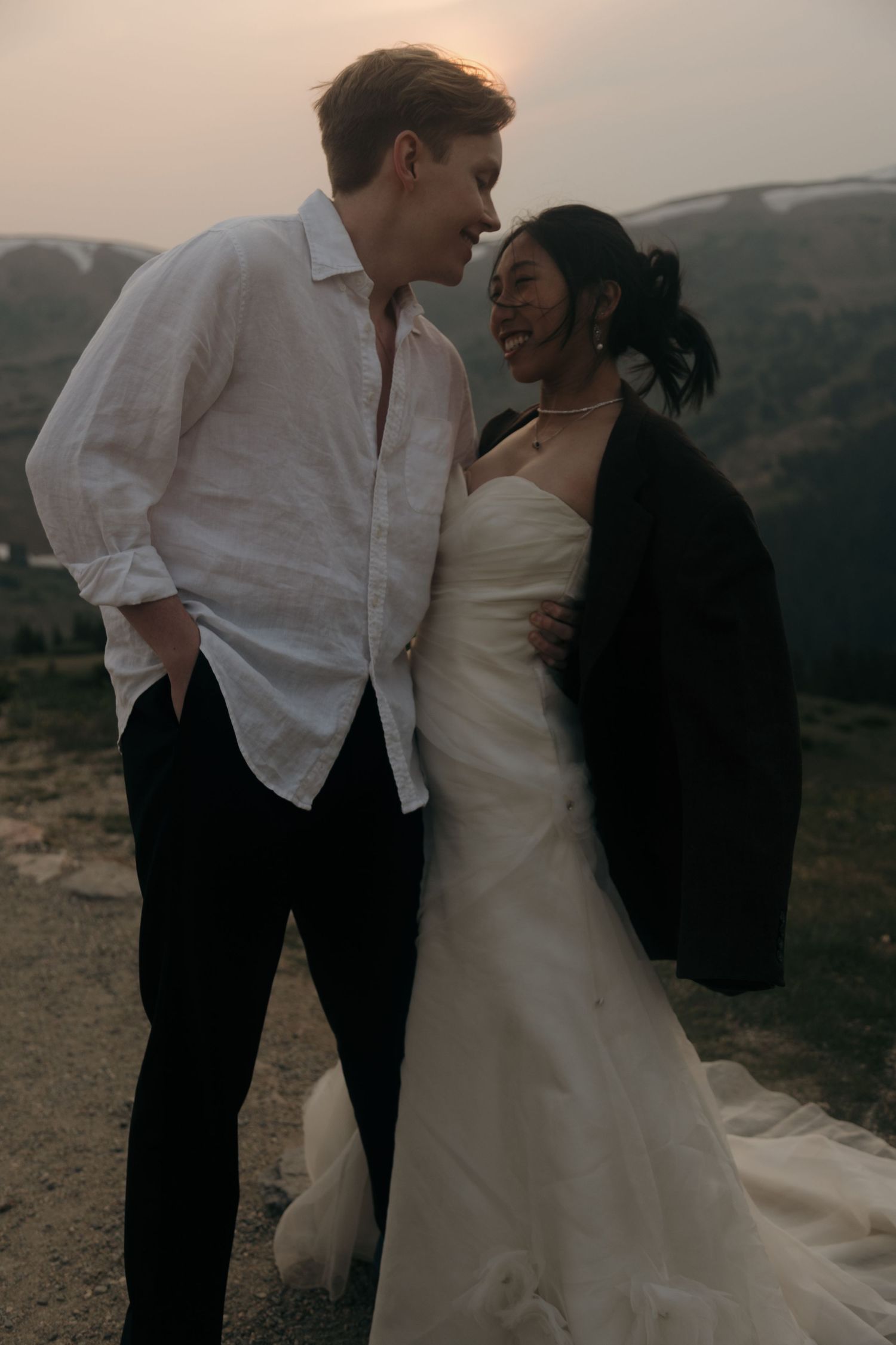Wedding couple embracing at sunset on a mountain overlook, bride wearing groom's dark jacket.