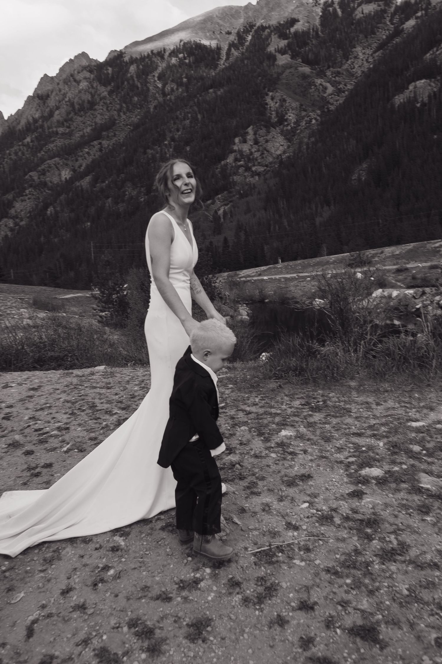 Laughing bride in white gown holds hands with young child in mountain setting.