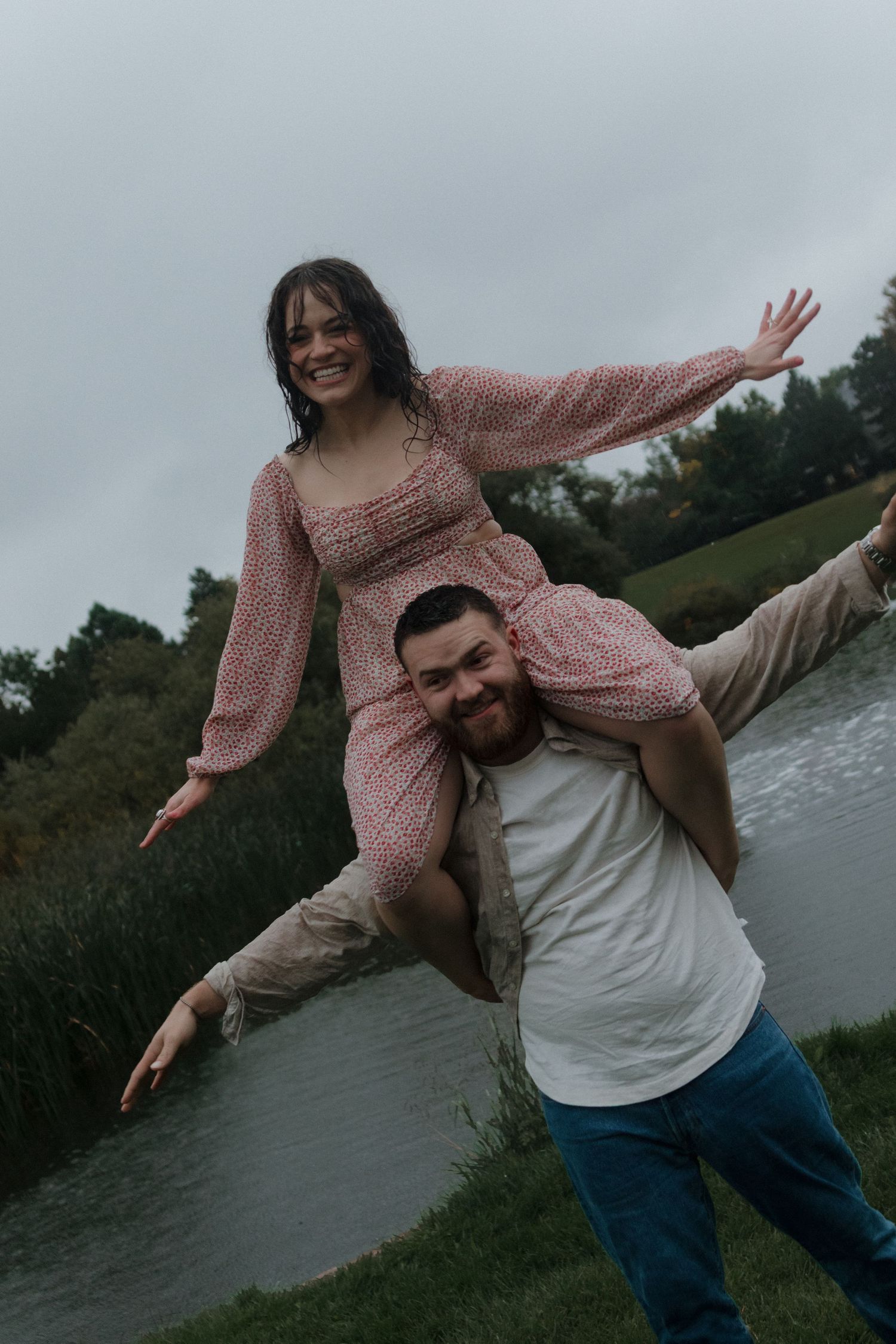 Playful couple posing by a lake, with one person being carried while spreading their arms.