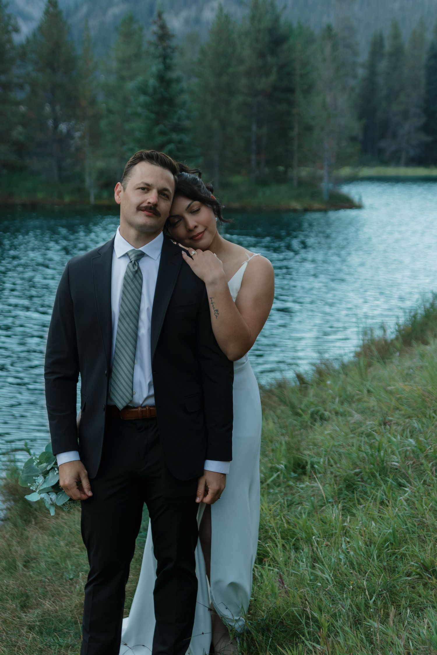 A romantic lakeside wedding portrait with mountains and evergreen trees in the background.