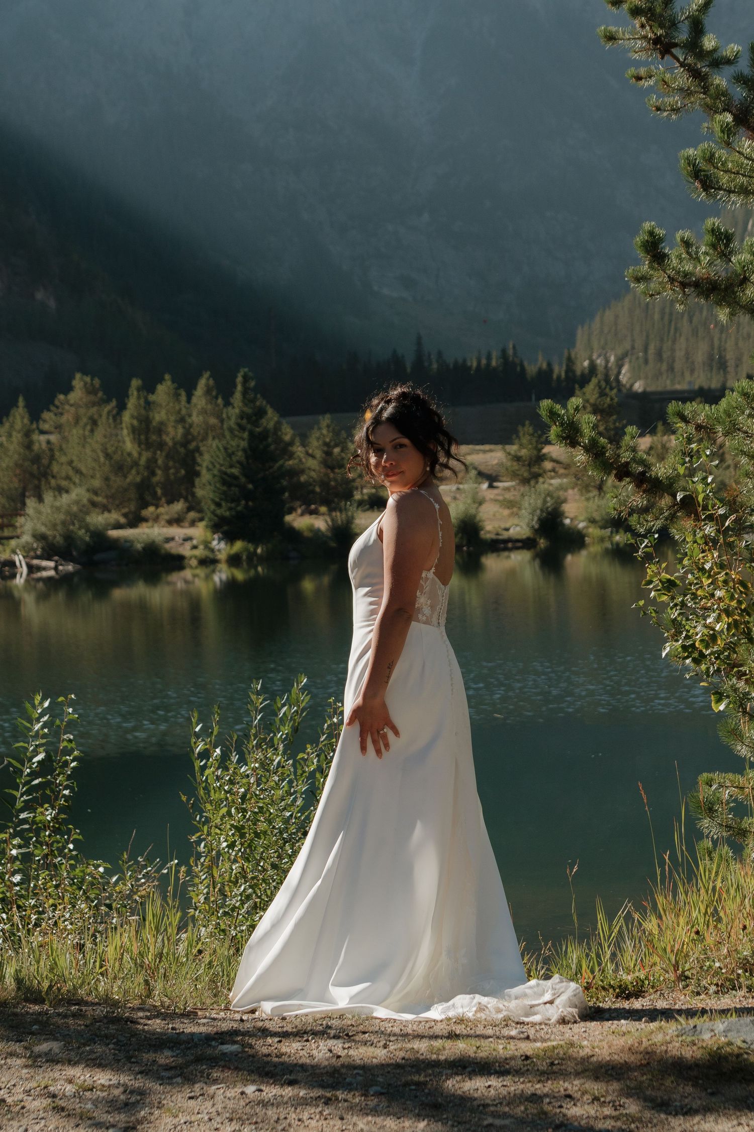 A bride in a flowing white dress stands against a scenic mountain lake backdrop.