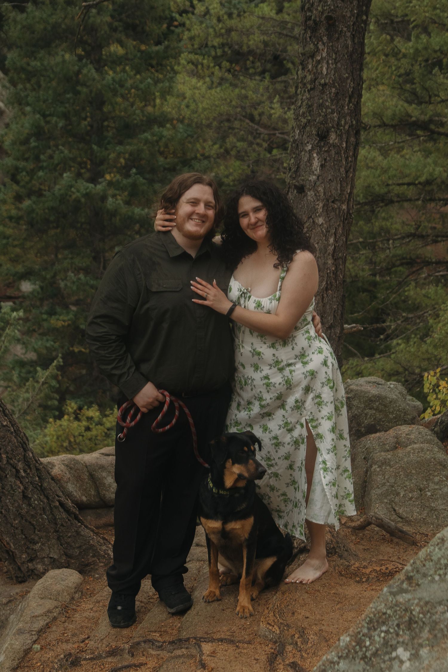 A couple and their rottweiler puppy pose together on a rocky overlook in a scenic forest setting.