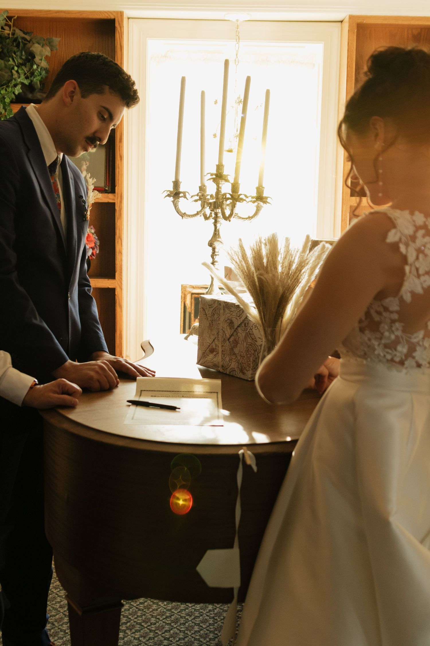 Couple signs documents at a wooden table with candlesticks in warm indoor lighting.