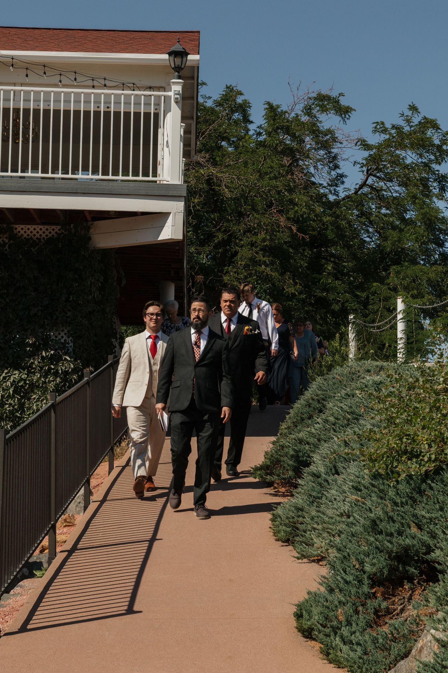 Groomsmen in formal attire walking along curved garden pathway.