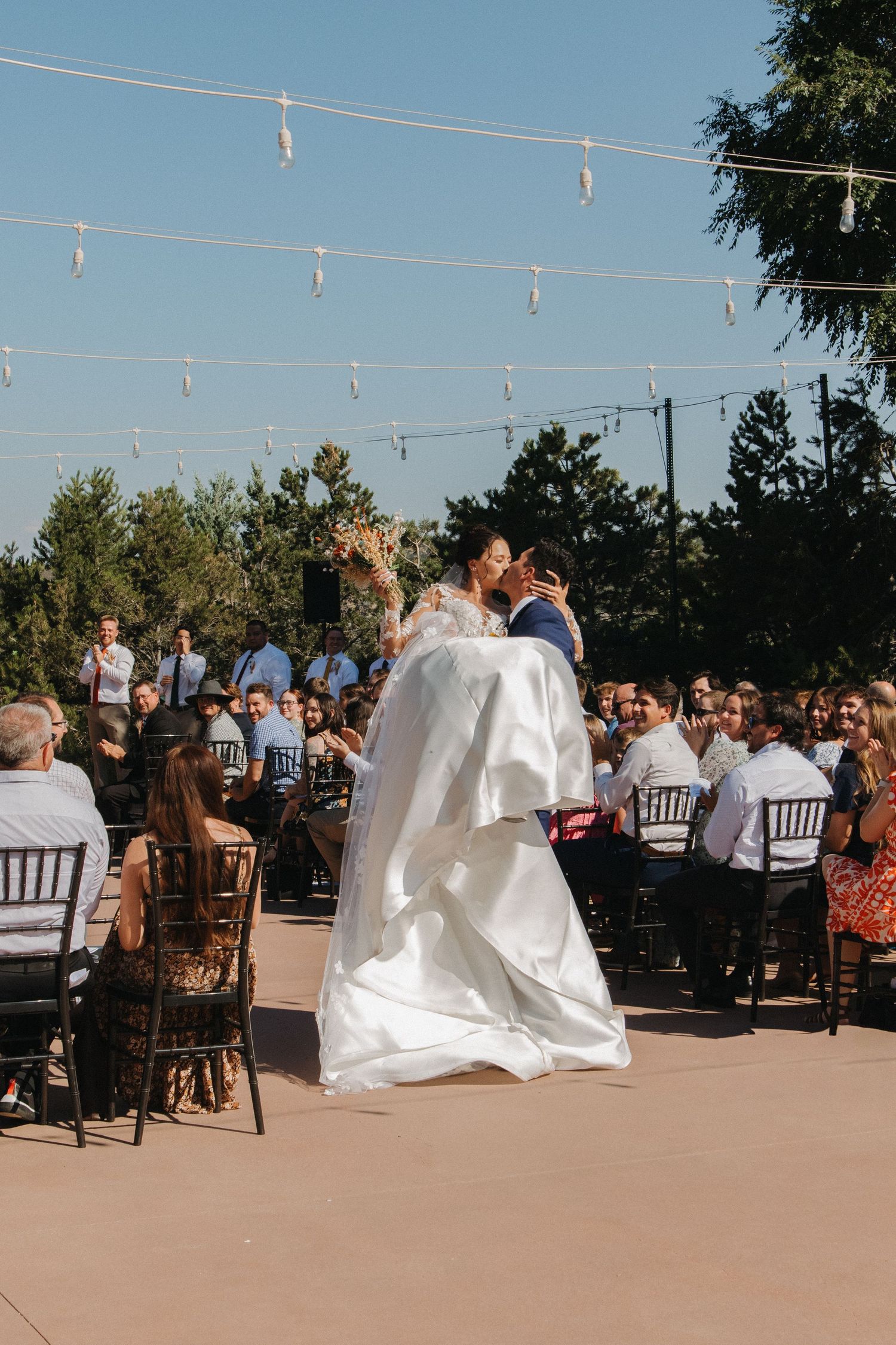 Outdoor wedding ceremony with string lights and guests seated in wooden chairs.