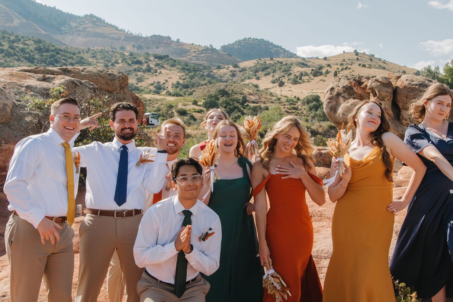 Wedding party members in mixed formal attire pose against mountain landscape.