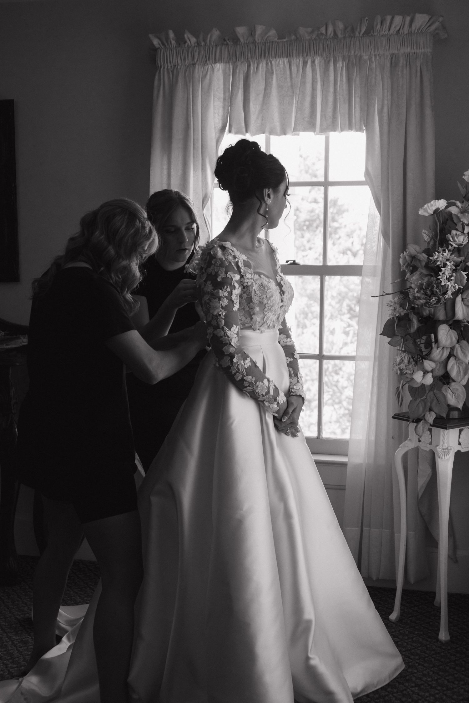 Black and white photo capturing final wedding dress adjustments in a sunlit room with elegant floral arrangements.