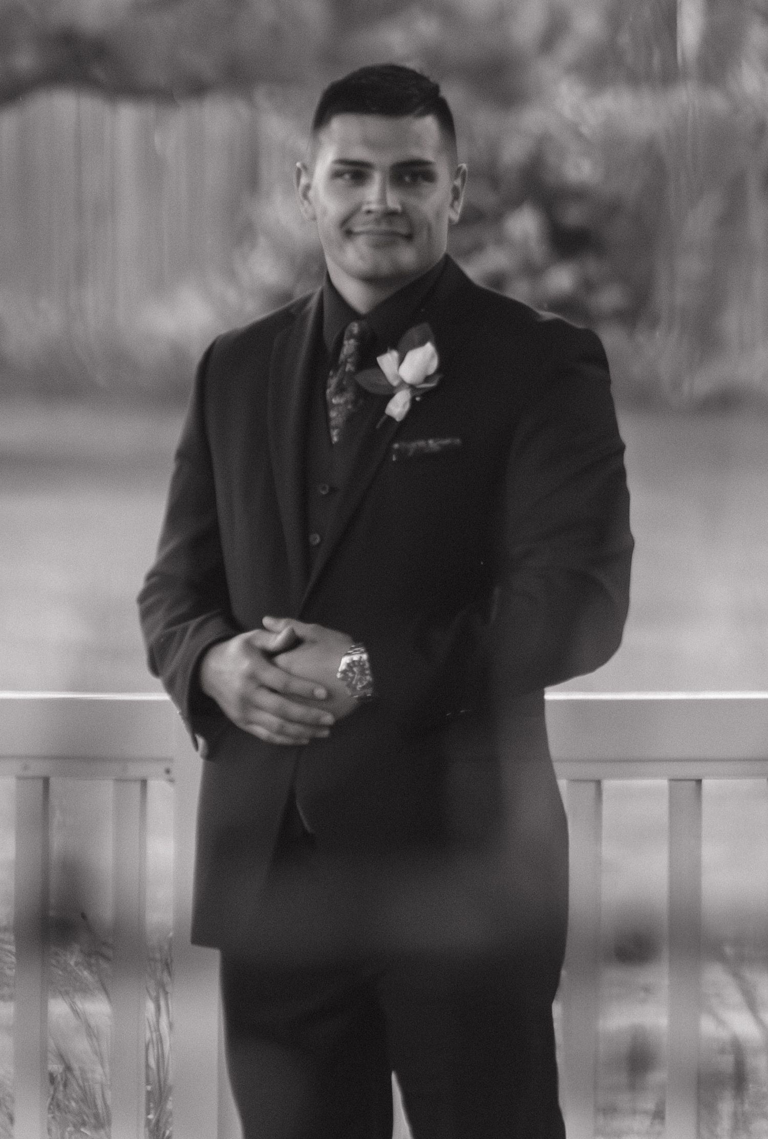 A formal portrait of a person in a black tuxedo with a boutonniere at a wedding event.