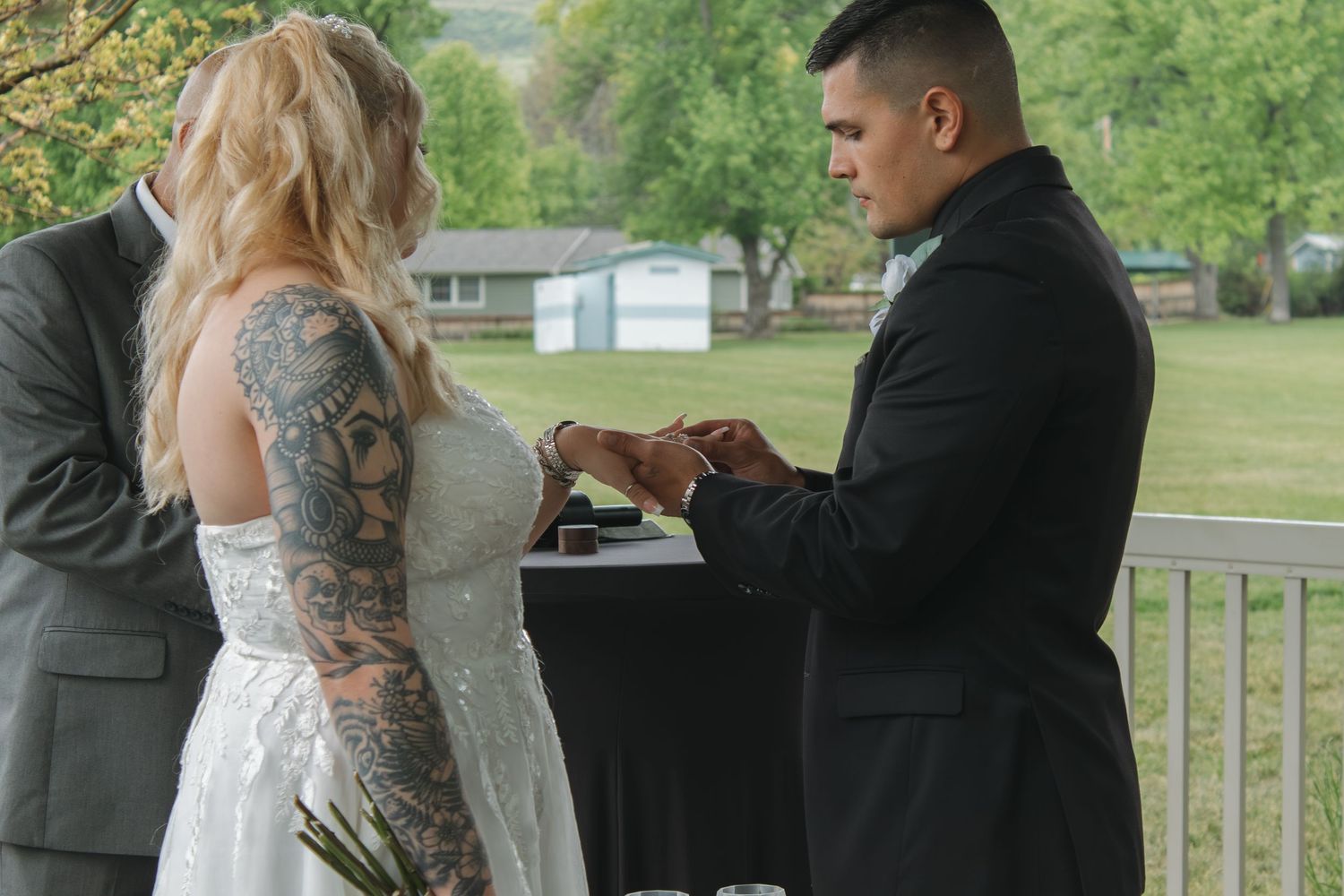 A wedding ceremony takes place in a green park pavilion with trees in the background.