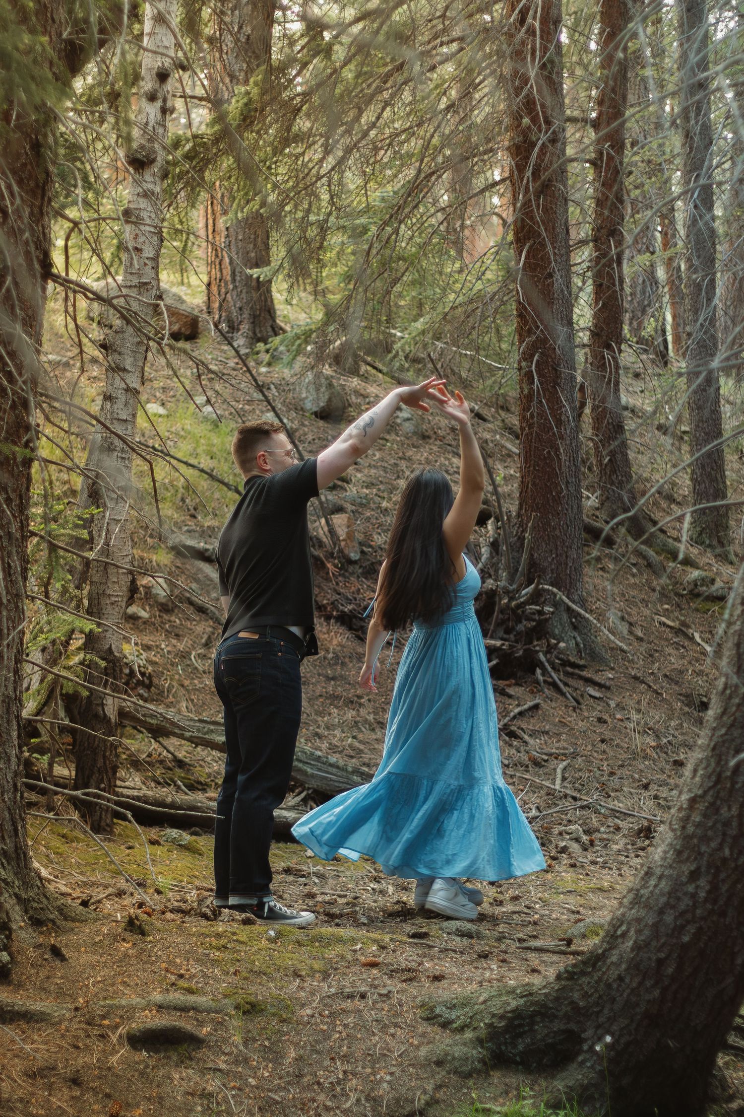 A couple dances together on a forest path, with the person in blue twirling in a flowing dress.