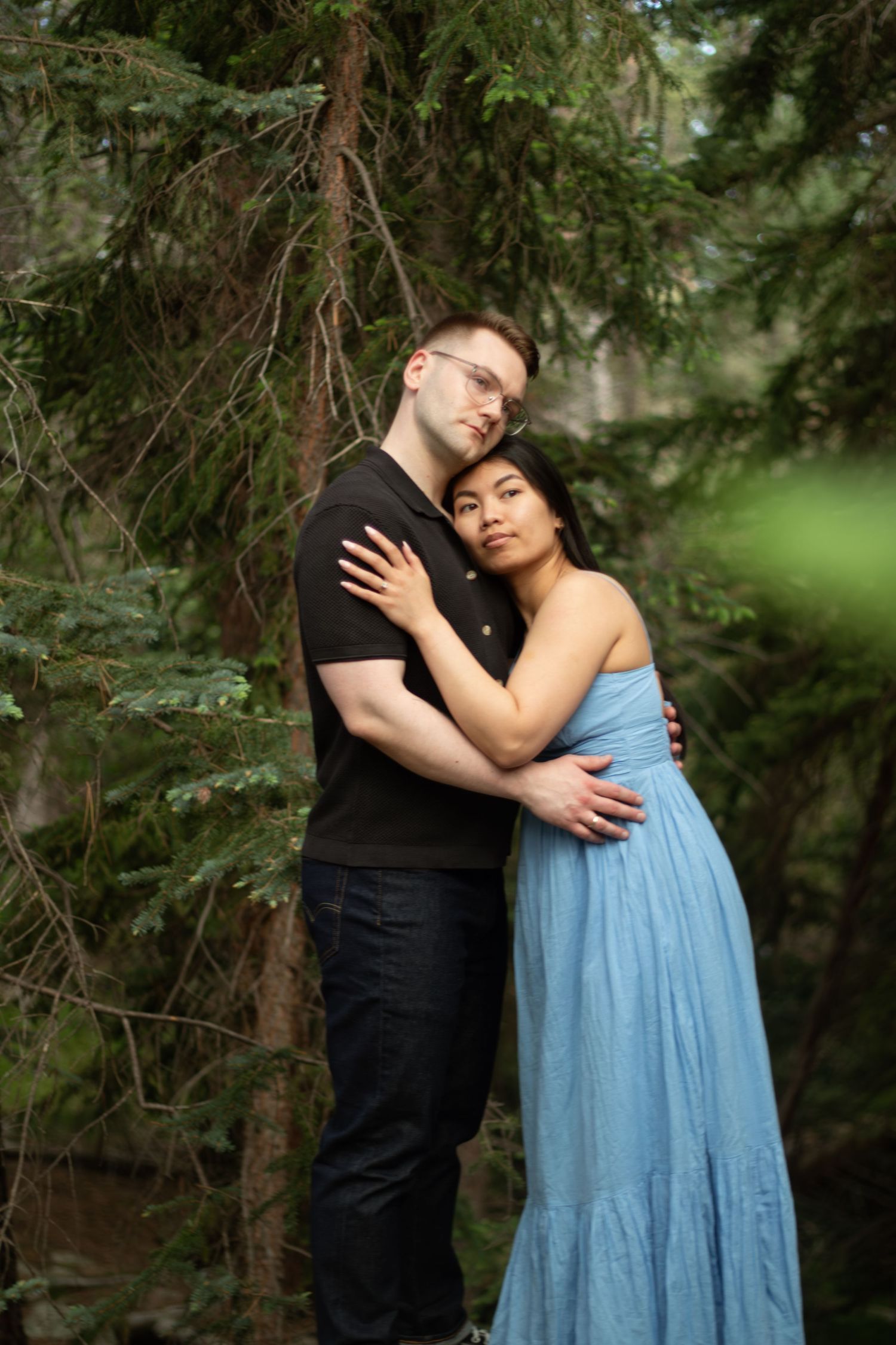 A tender embrace between two people surrounded by evergreen branches in a moody forest setting.