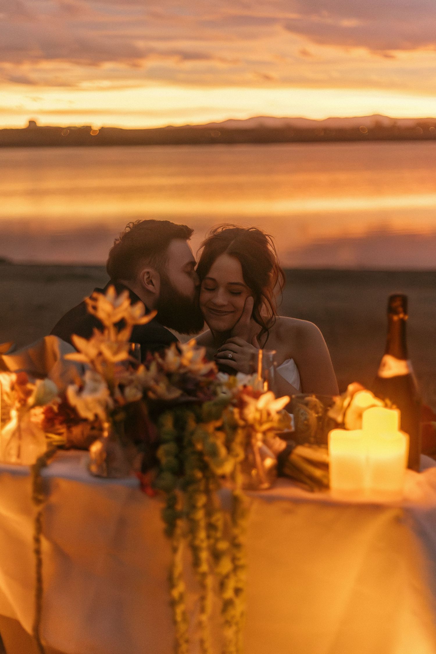 Candlelit wedding reception table decorated with flowers against a stunning orange and pink sunset backdrop.