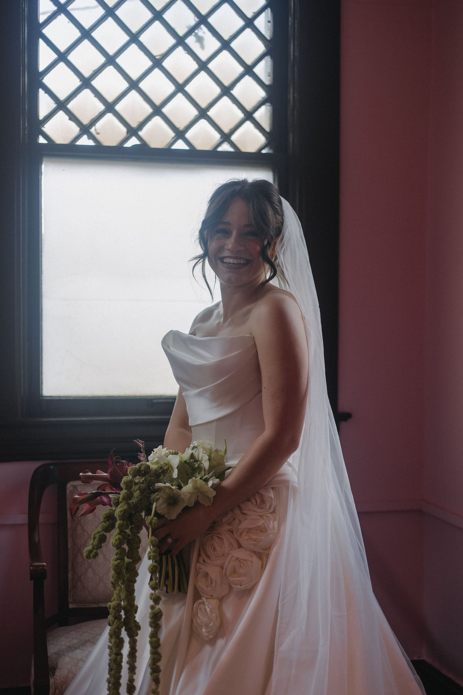 Bride in flowing white dress posing by latticed windows in a room with red walls.