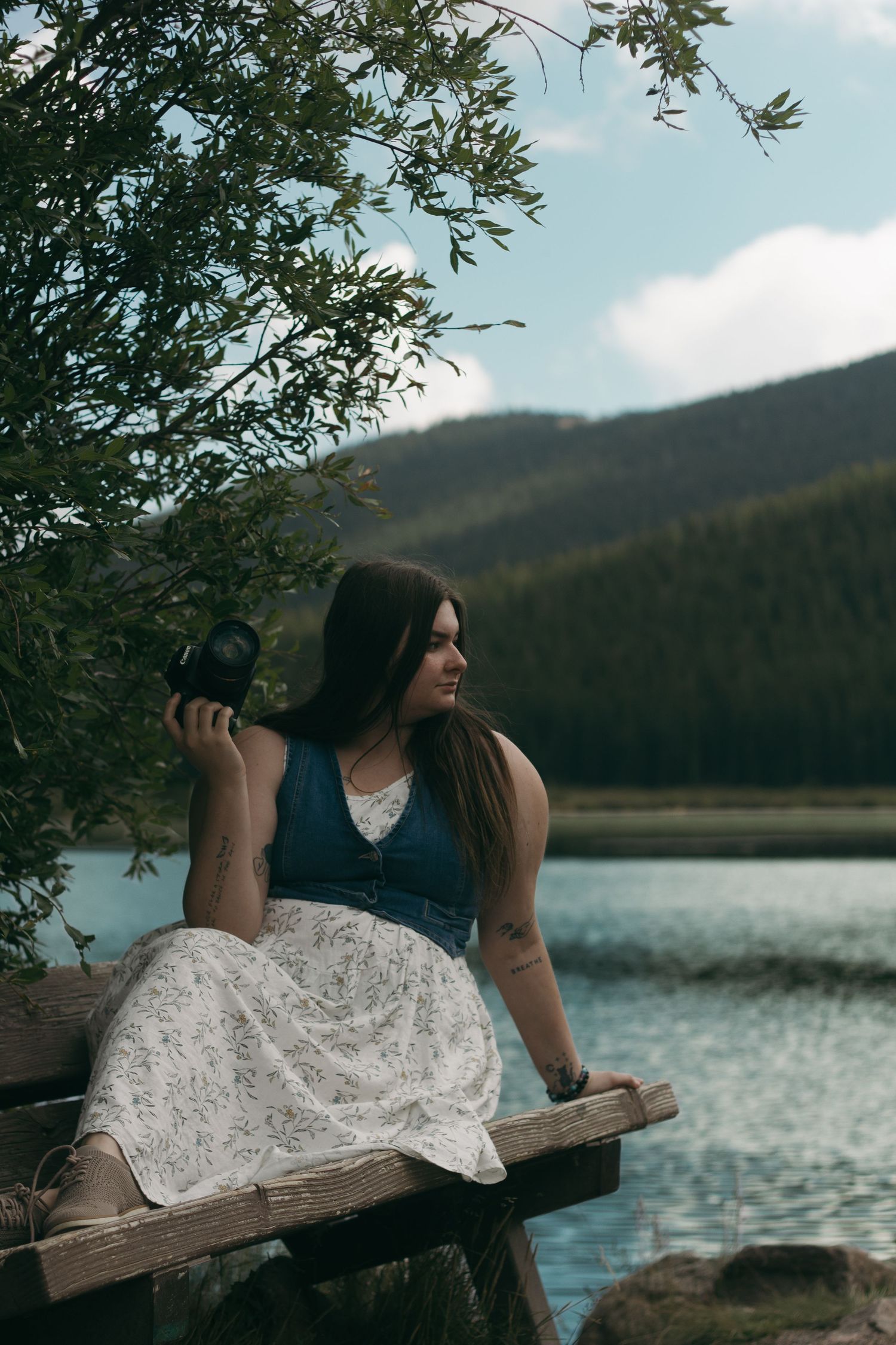 A person in a black top and white dress reclines on a wooden bench by a serene mountain lake at sunset.