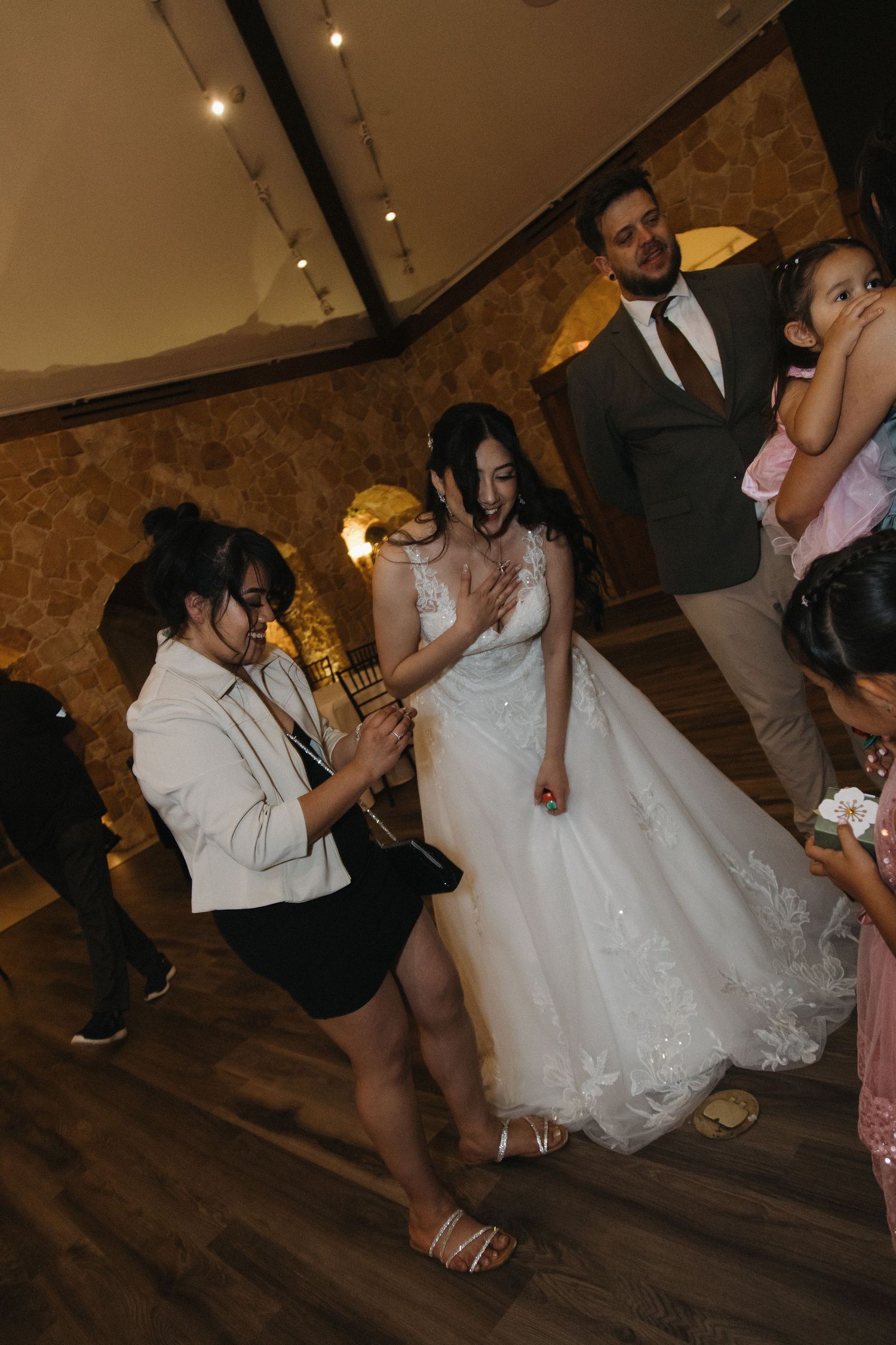 Bride in lace gown laughing with guests inside a warmly lit rustic stone wedding reception hall.