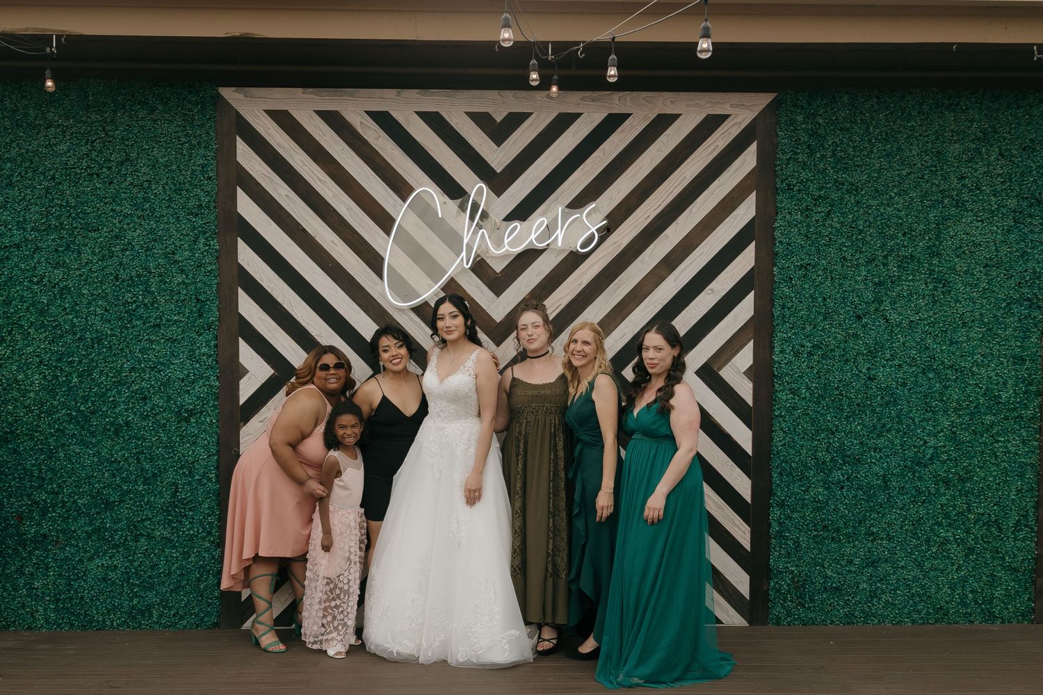 Bride in white gown poses with five bridesmaids in colorful dresses in front of geometric Cheers neon sign backdrop.