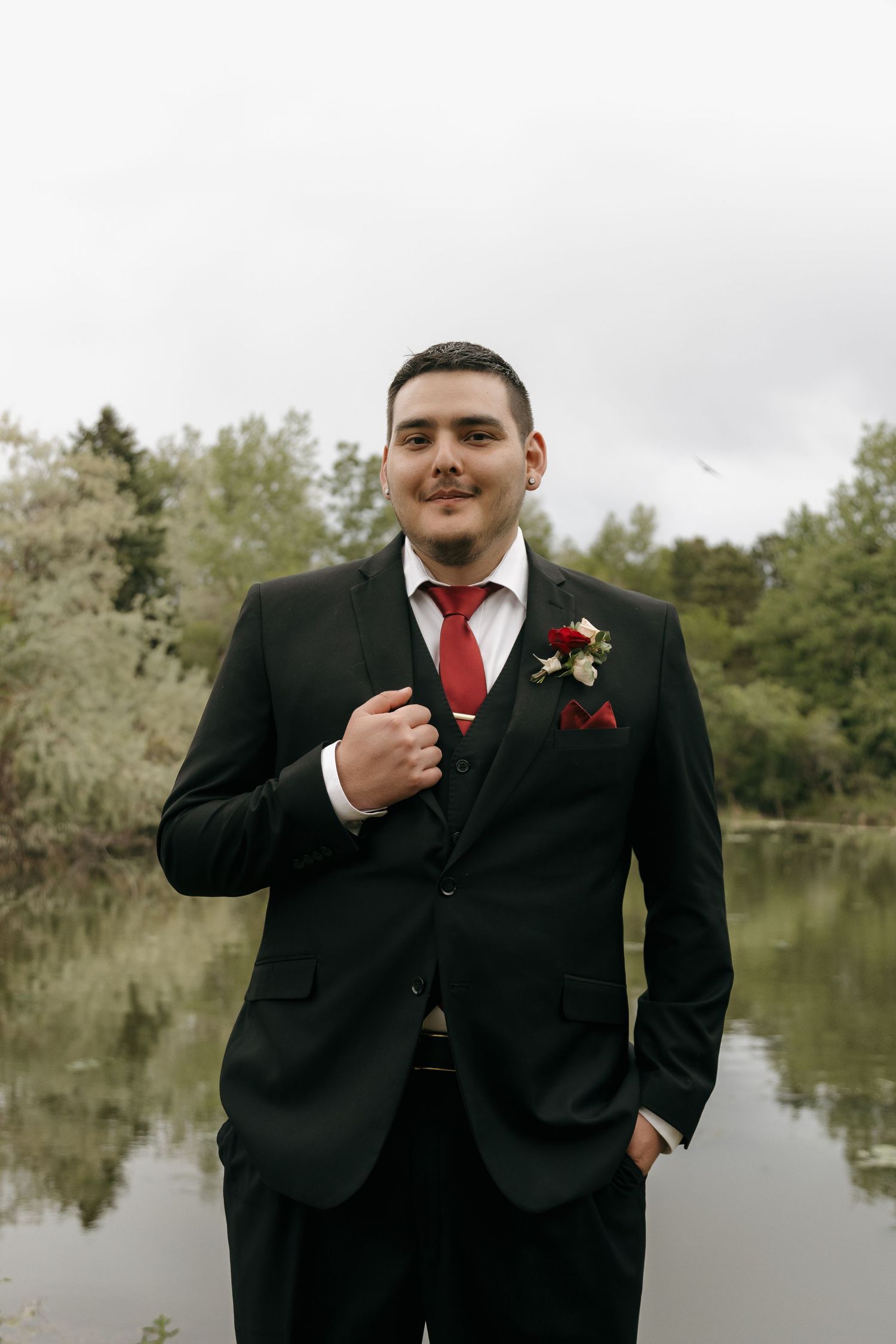 Dapper groom in black suit with red tie and floral boutonniere posing near a lake.
