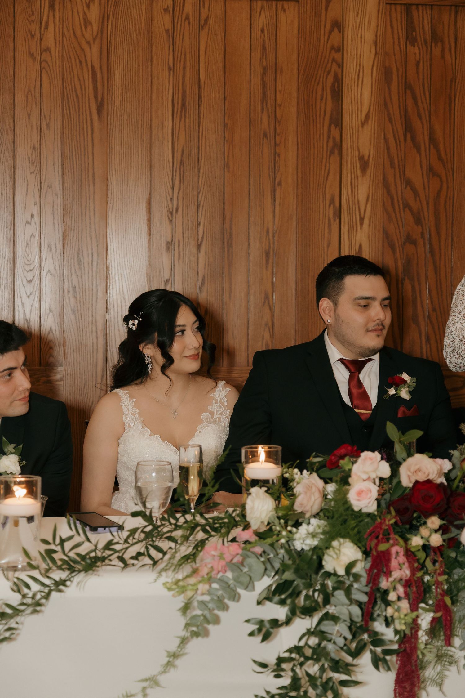 Bride and groom seated at sweetheart table adorned with lush floral arrangements of red and pink roses.