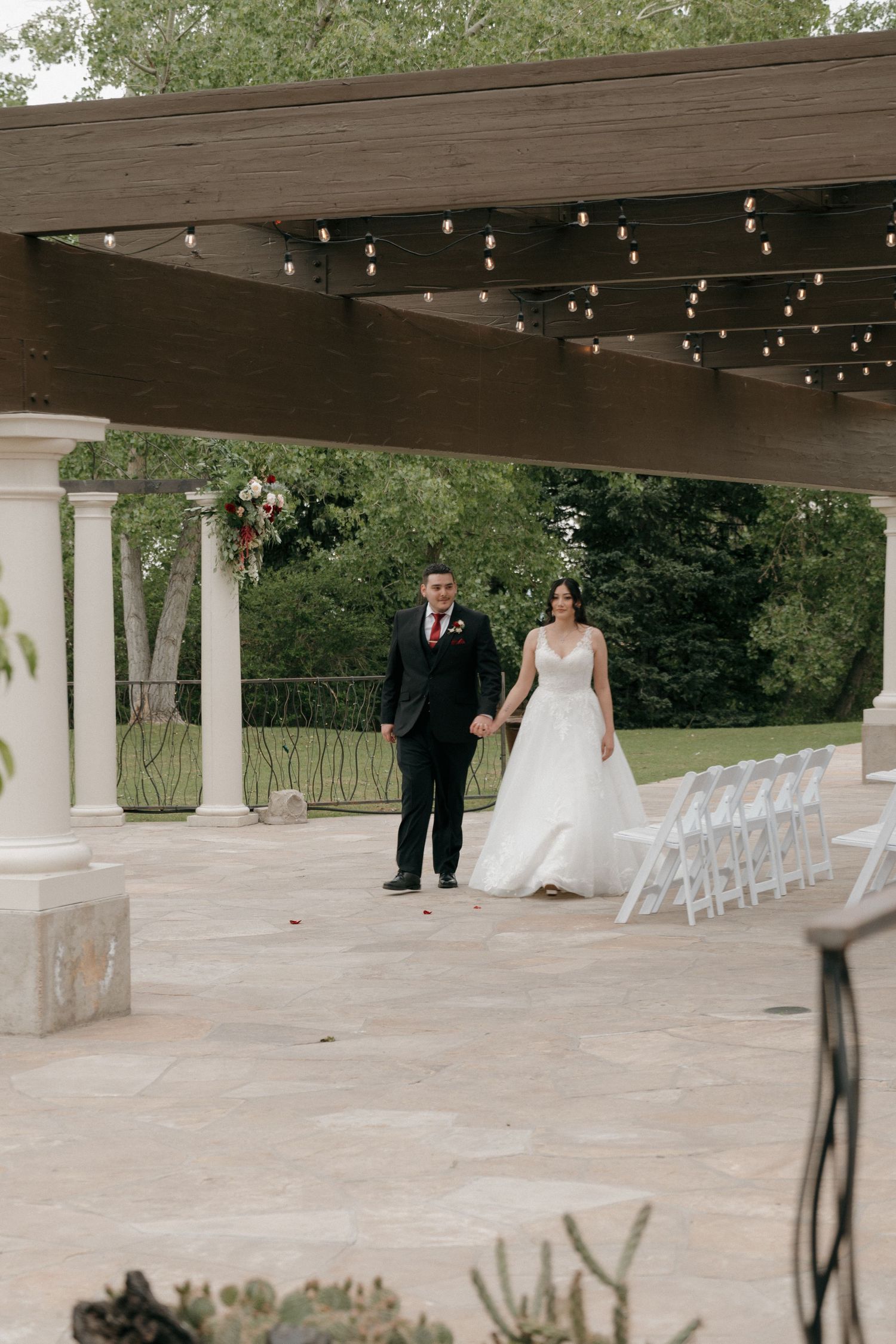 Bride and groom walk hand-in-hand along a stone pathway beneath a pergola with string lights at their venue.