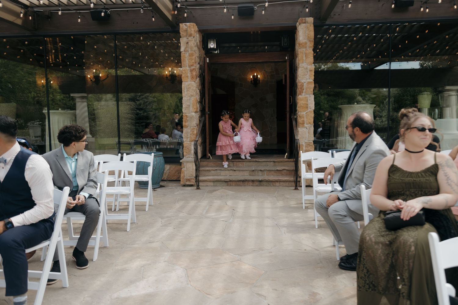 Flower girls in pink dresses walk down outdoor wedding aisle lined with white chairs and guests.