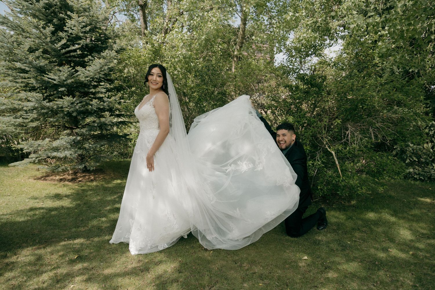 Groom kneels to spread bride's elegant white wedding gown train in a lush garden setting.