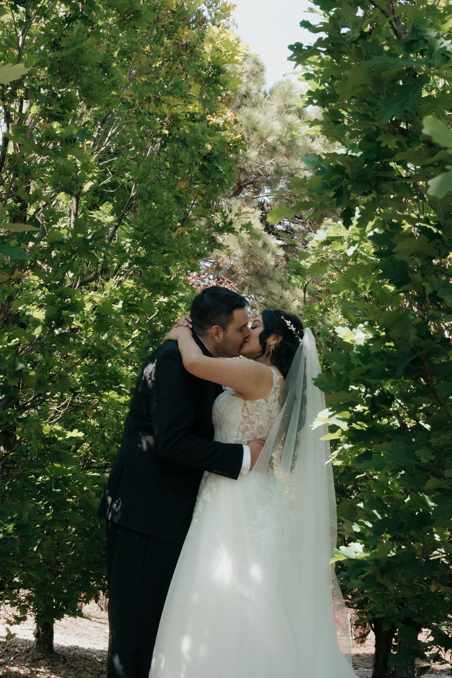 Bride and groom share a romantic kiss surrounded by lush green trees during their outdoor wedding ceremony.