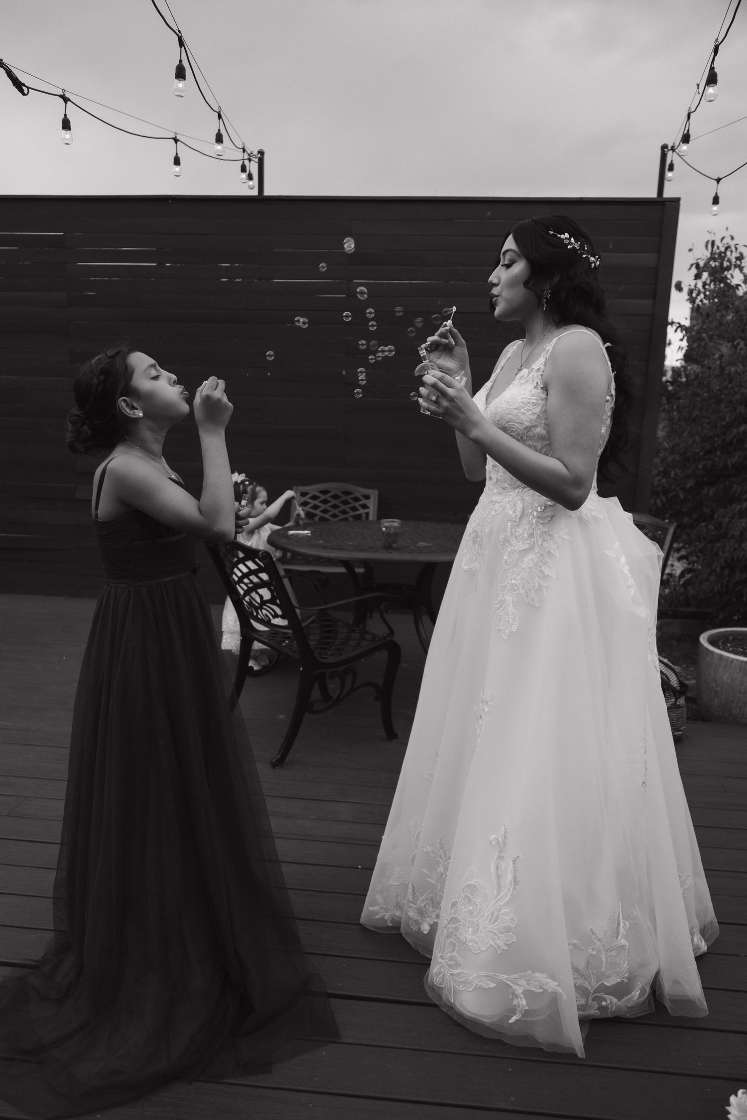 Bride in white lace gown blows bubbles with young bridesmaid on outdoor deck in black and white photo.