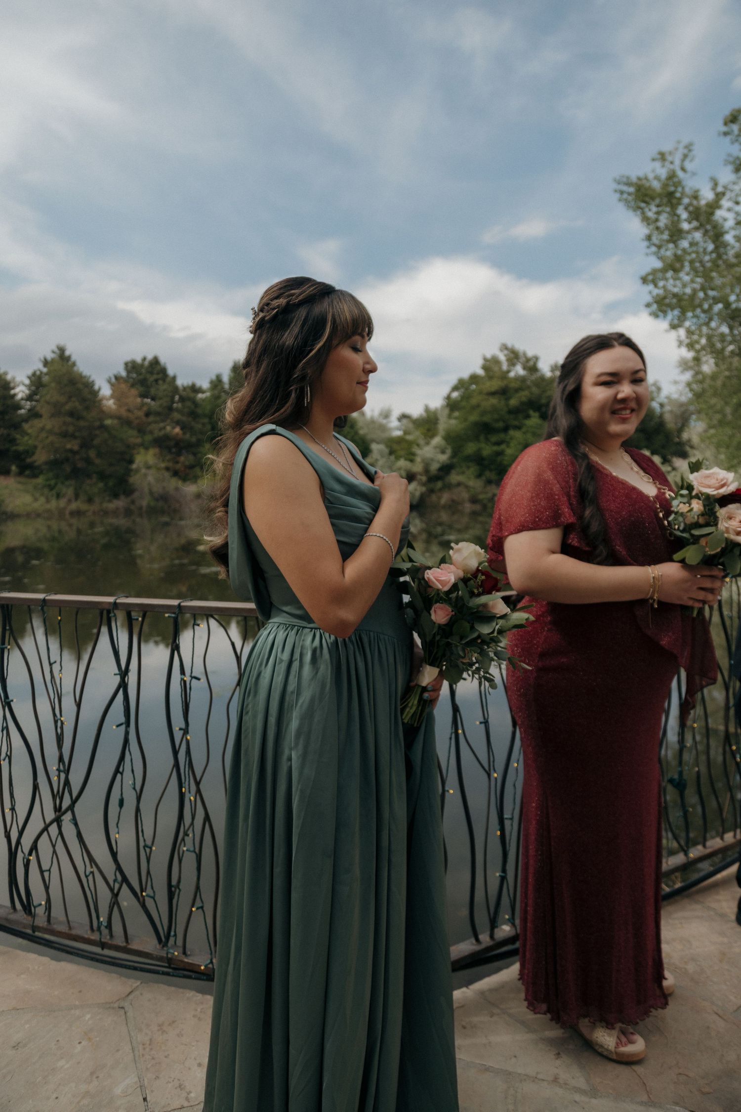 Two bridesmaids in green and burgundy gowns stand on a bridge overlooking a serene lake outdoors.