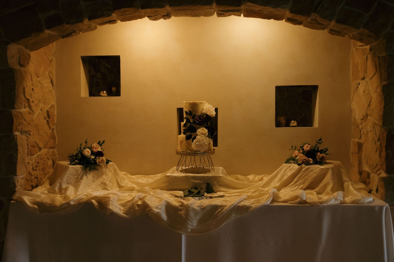 Elegant wedding cake display on draped table in stone arch alcove with floral arrangements and warm lighting.