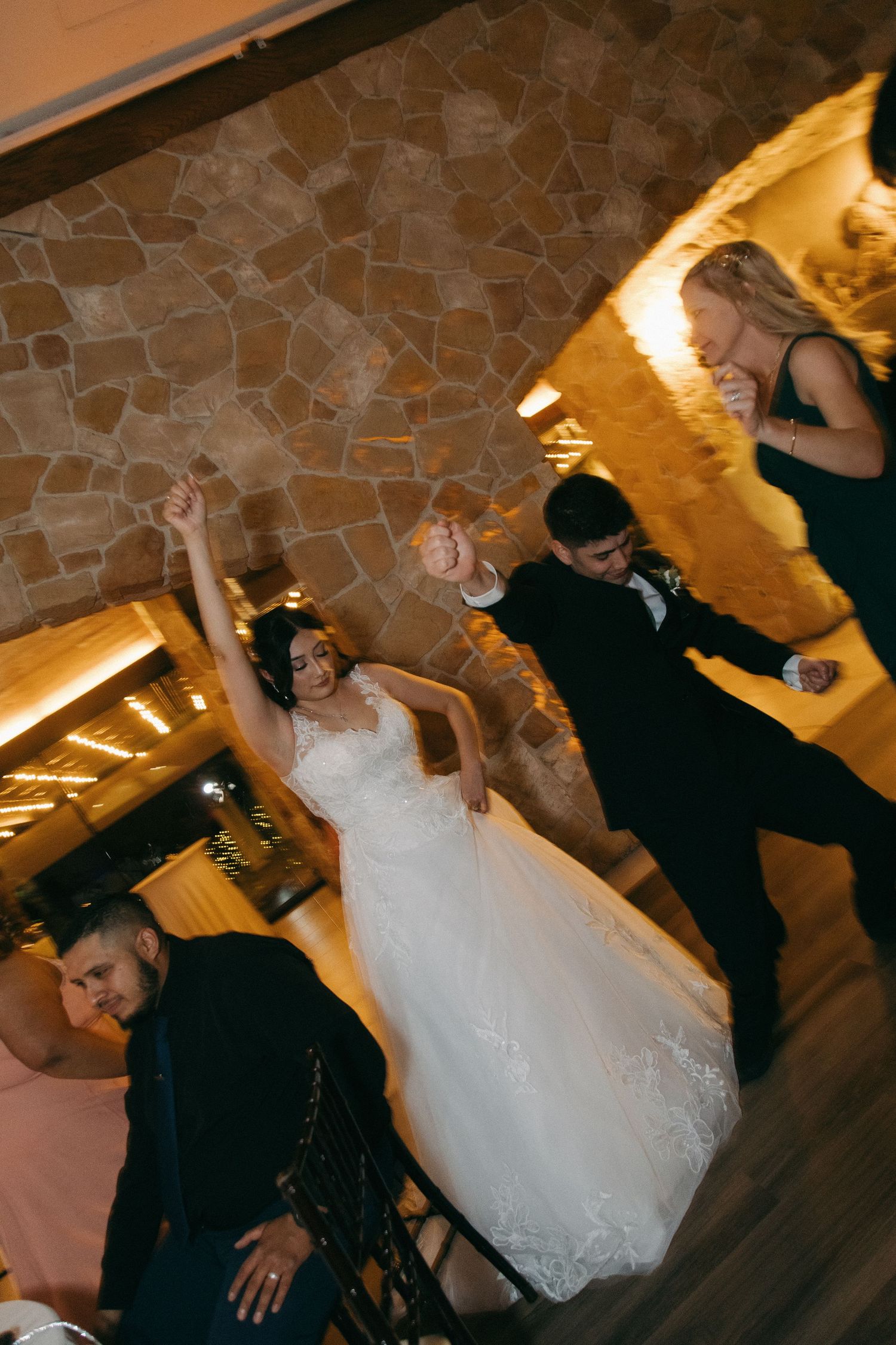 Bride raises her arms joyfully while dancing with guests at her wedding reception near stone walls.