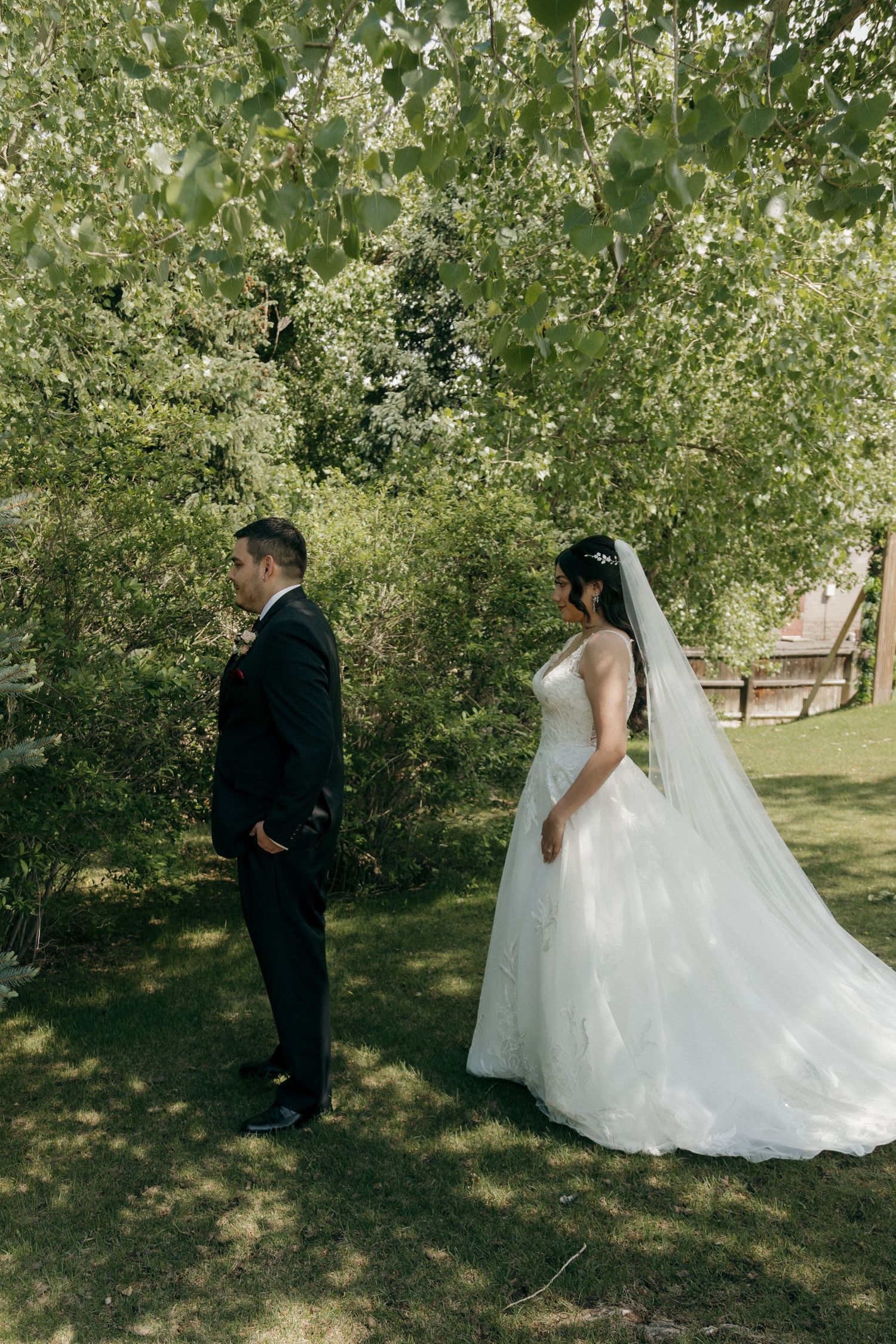Bride and groom standing back to back among lush green trees during first look wedding photos.