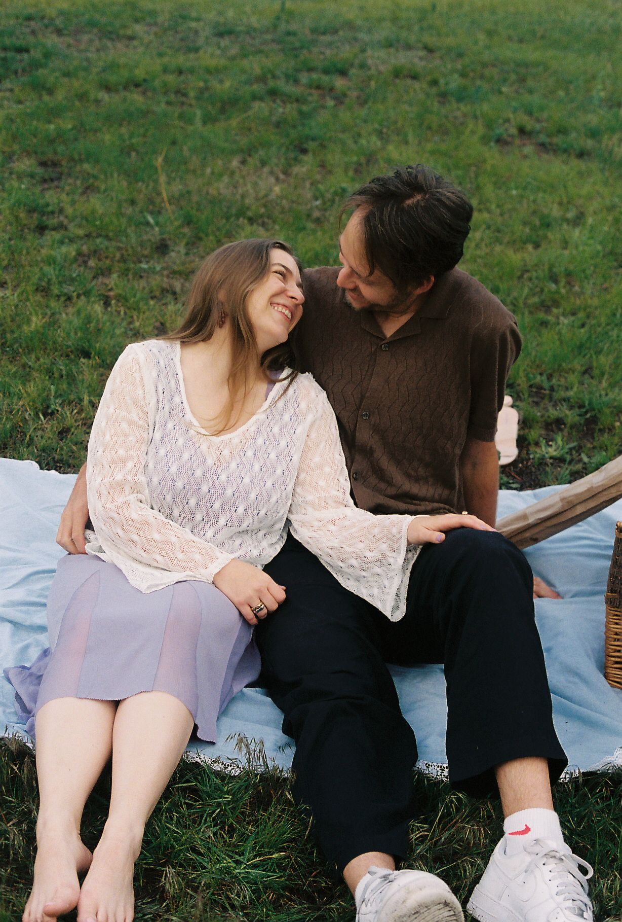 Couple sitting on a blanket in a green field, smiling and gazing at each other during a romantic picnic.