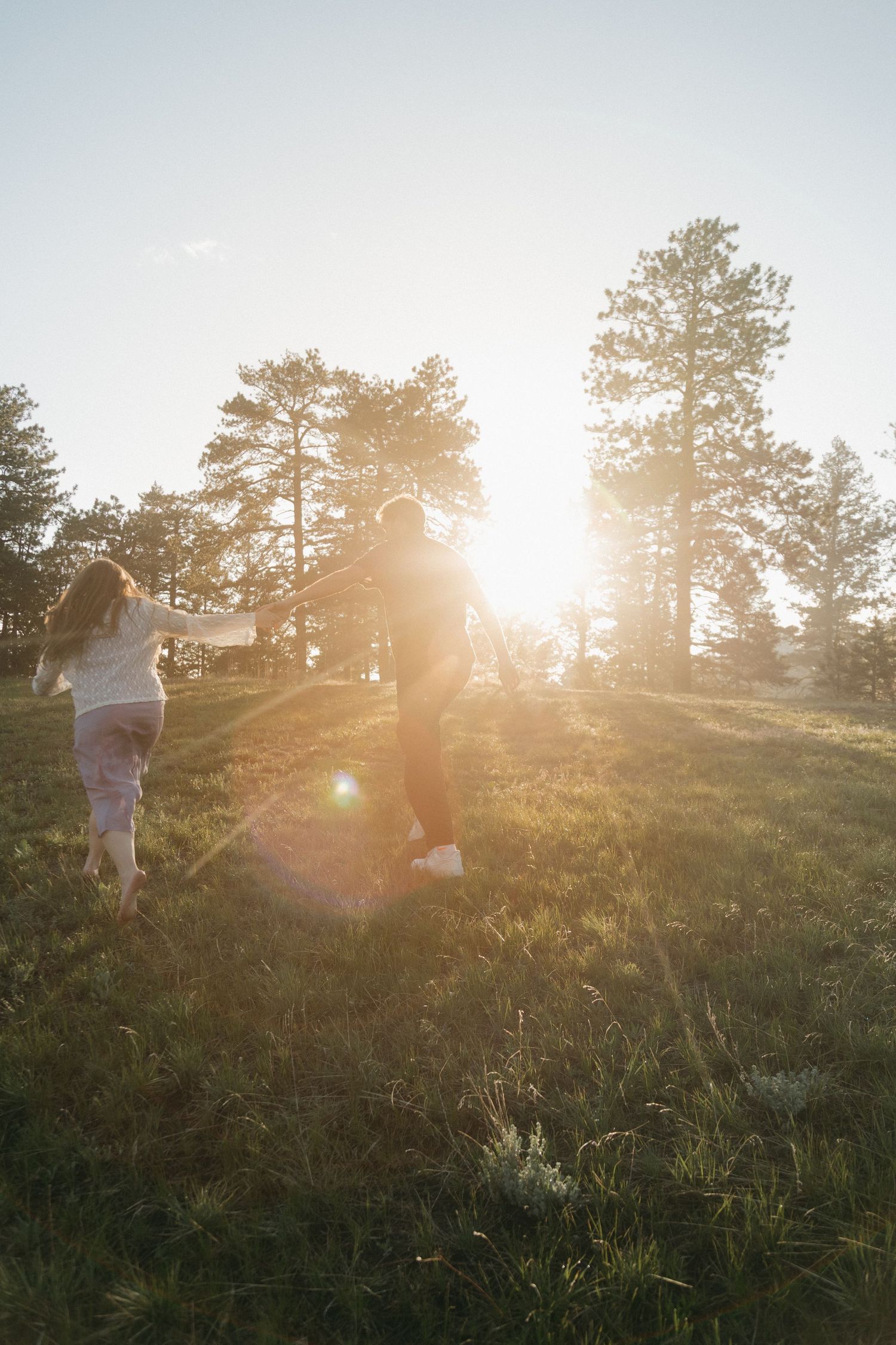 Couple running through a sunlit grassy field surrounded by tall pine trees at golden hour.