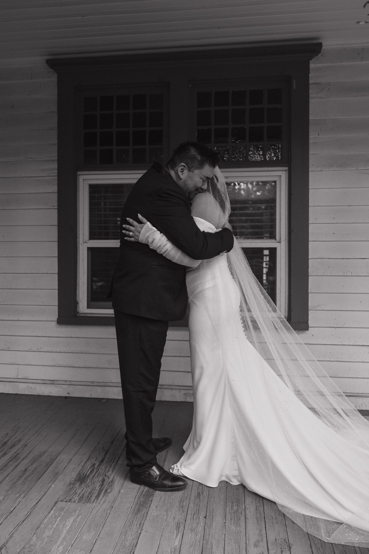 Black and white photo of bride and groom sharing tender embrace on wooden porch after wedding ceremony.